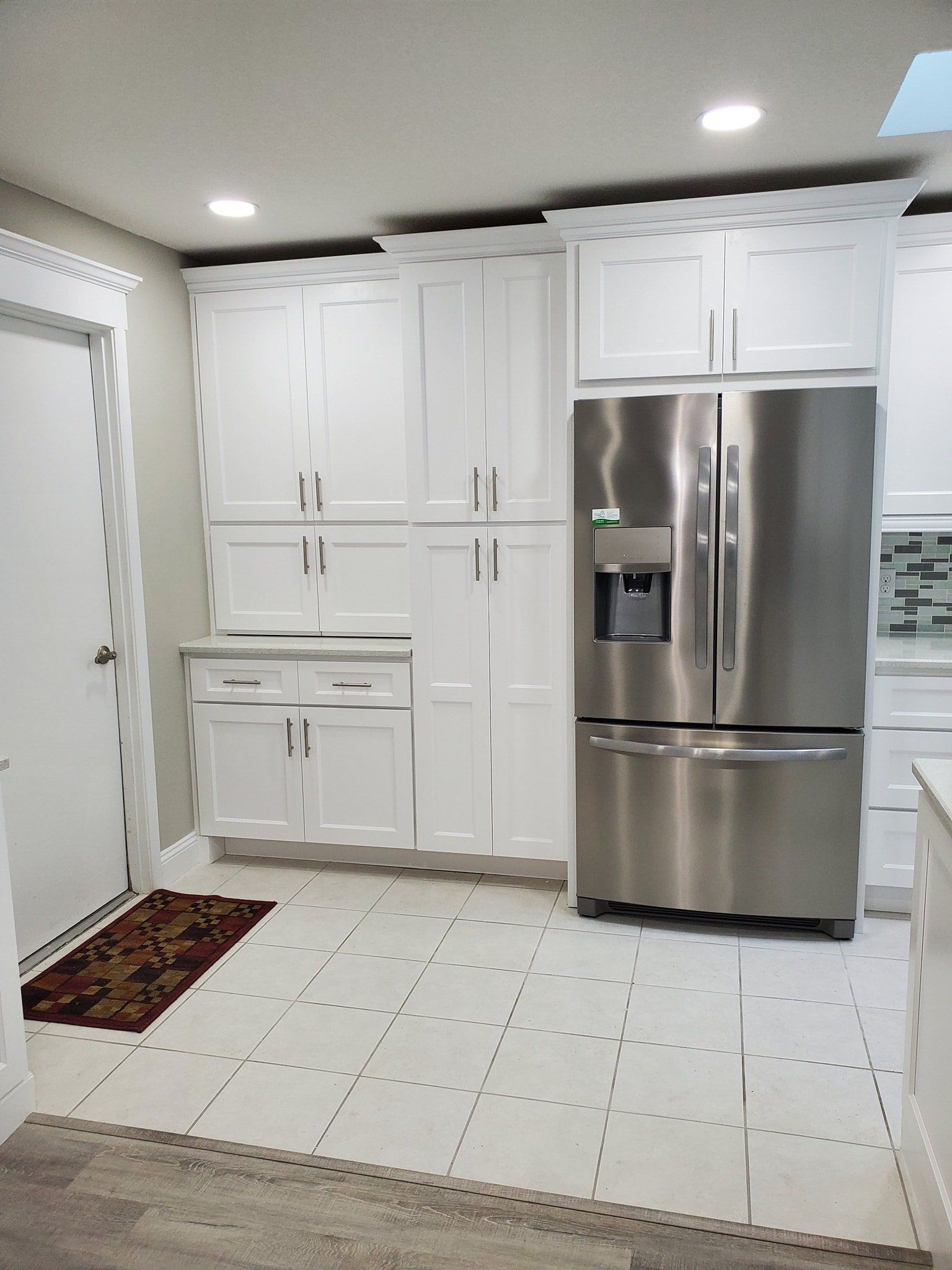 A white kitchen with tall cabinets, a stainless steel refrigerator, and tiled floors next to a door with a floor mat.