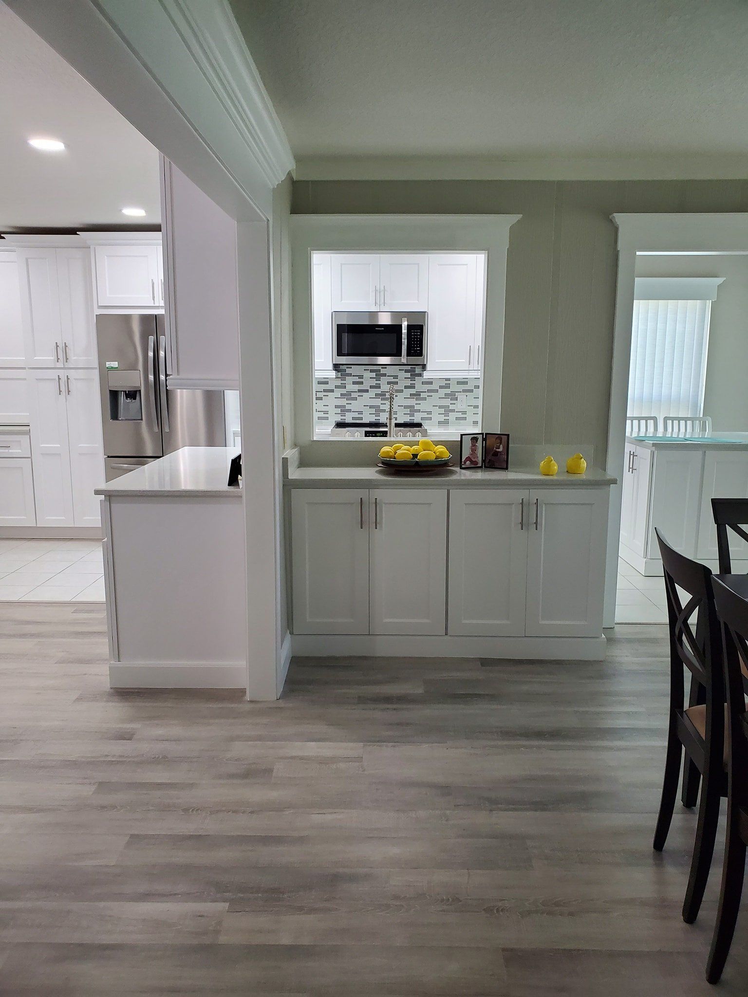 A view of a bright kitchen featuring white cabinets, wood-look flooring, and a serving window looking into a kitchen space.