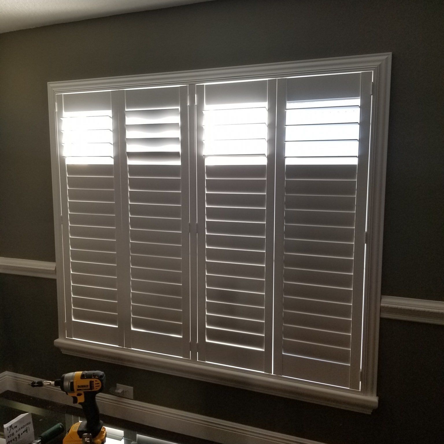 Four-panel white plantation shutters installed on a window in a room with dark walls and a drill on the windowsill.