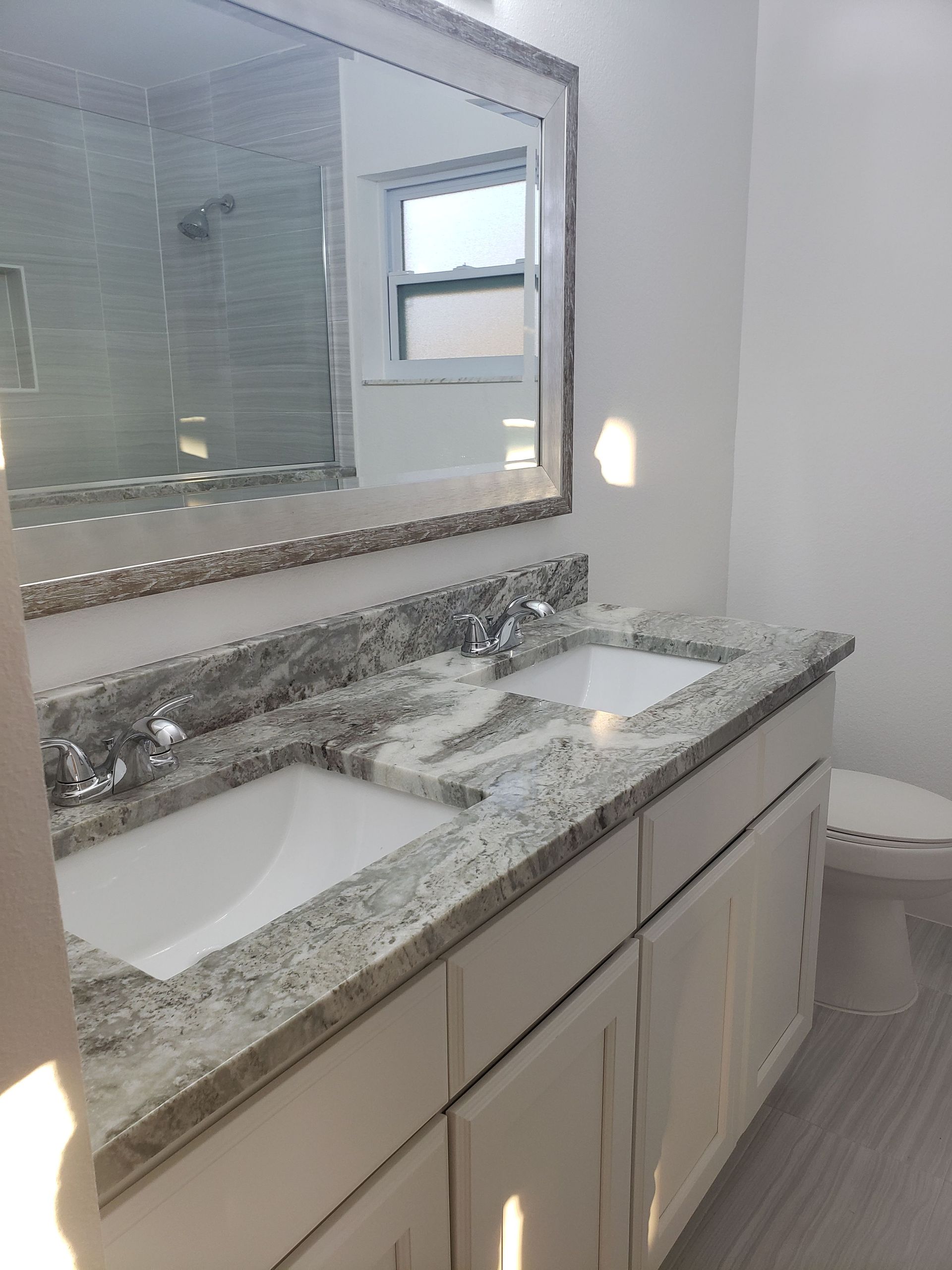 A bathroom vanity with a granite countertop, dual sinks, a large framed mirror, and a white cabinet, next to a toilet.