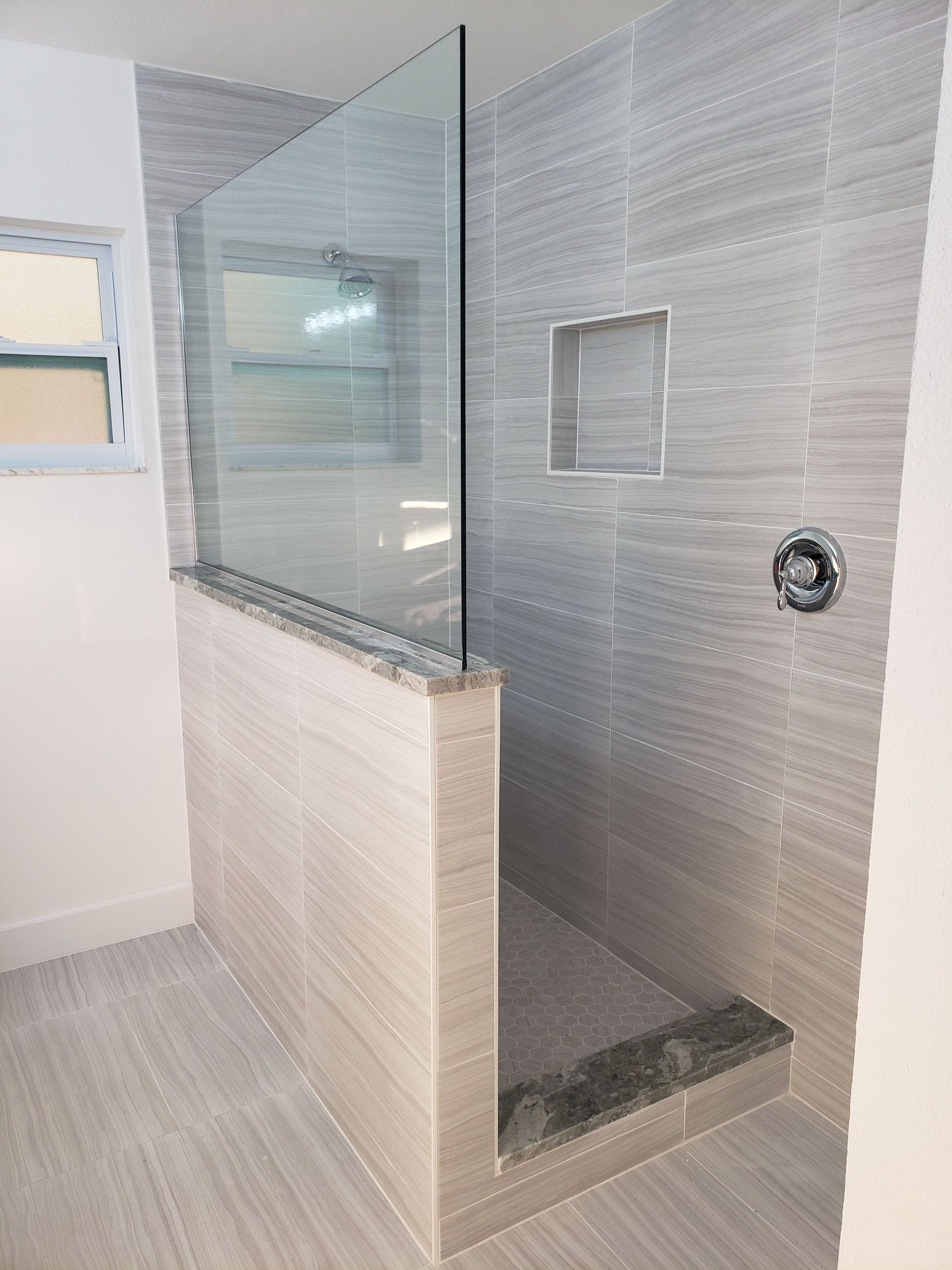 Modern walk-in shower with light grey tiled walls, a glass panel partition, a recessed shelf, and a dark stone threshold.