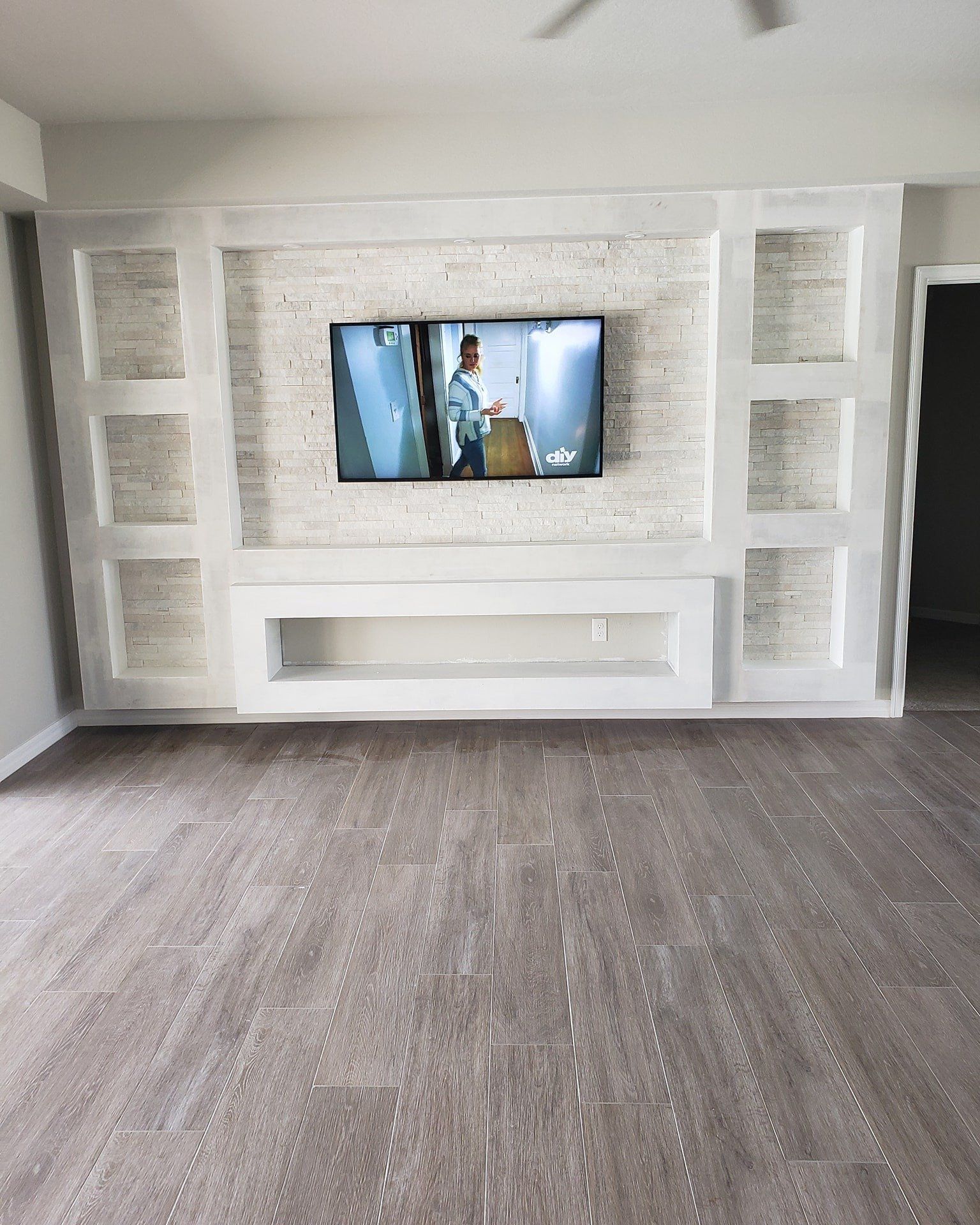 A wall-mounted TV centered on a light-colored stone accent wall with white built-in shelving, above a white fireplace niche.