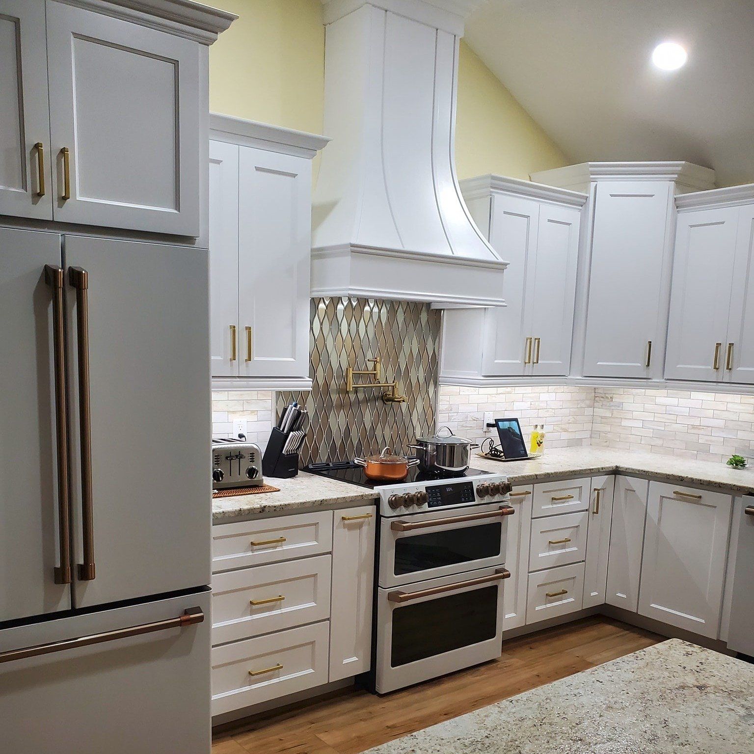 A modern kitchen with white shaker cabinets, gold hardware, a range, a vent hood, and a refrigerator.