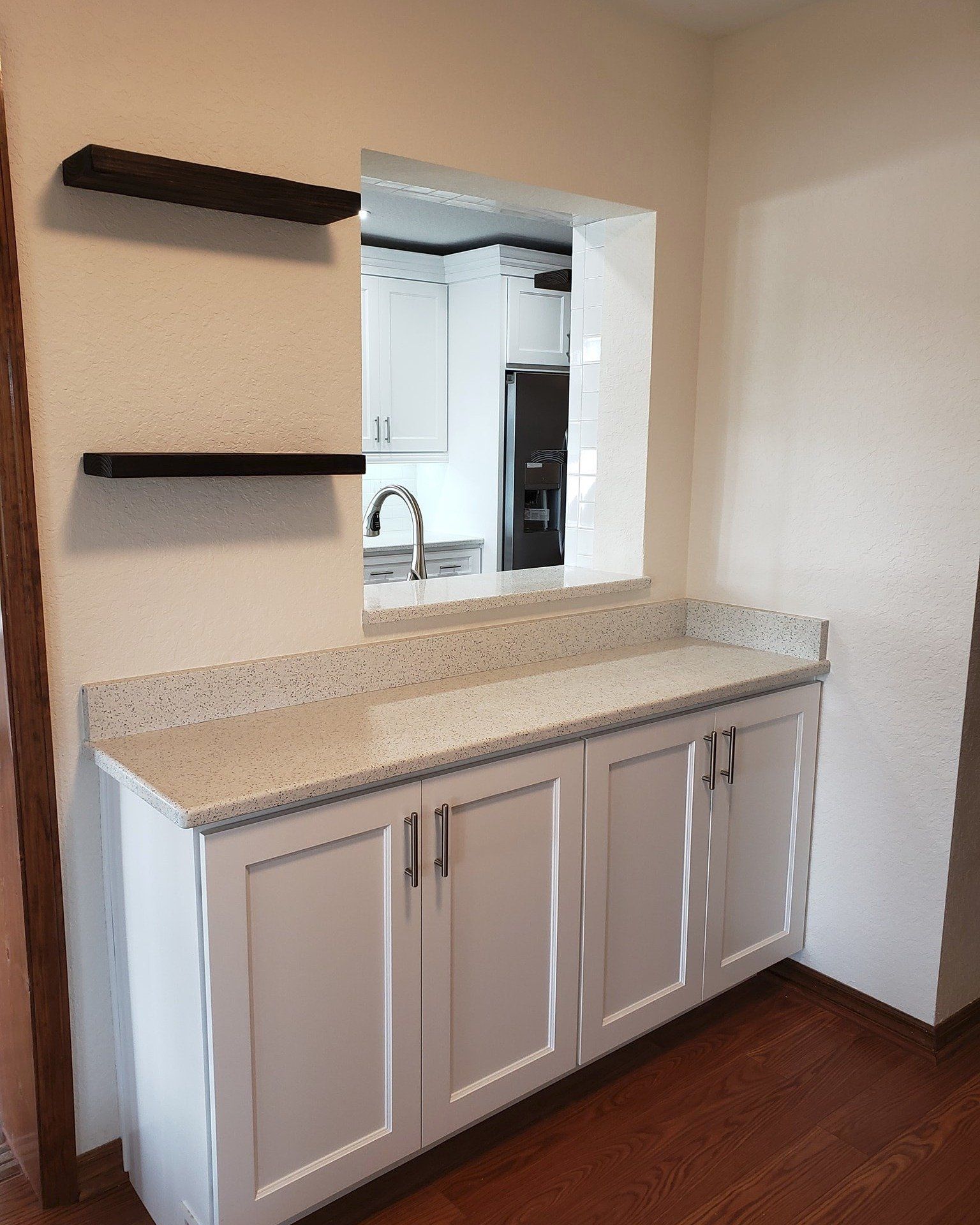 White cabinets with a speckled countertop and two black floating shelves mounted on the wall below a kitchen pass-through.