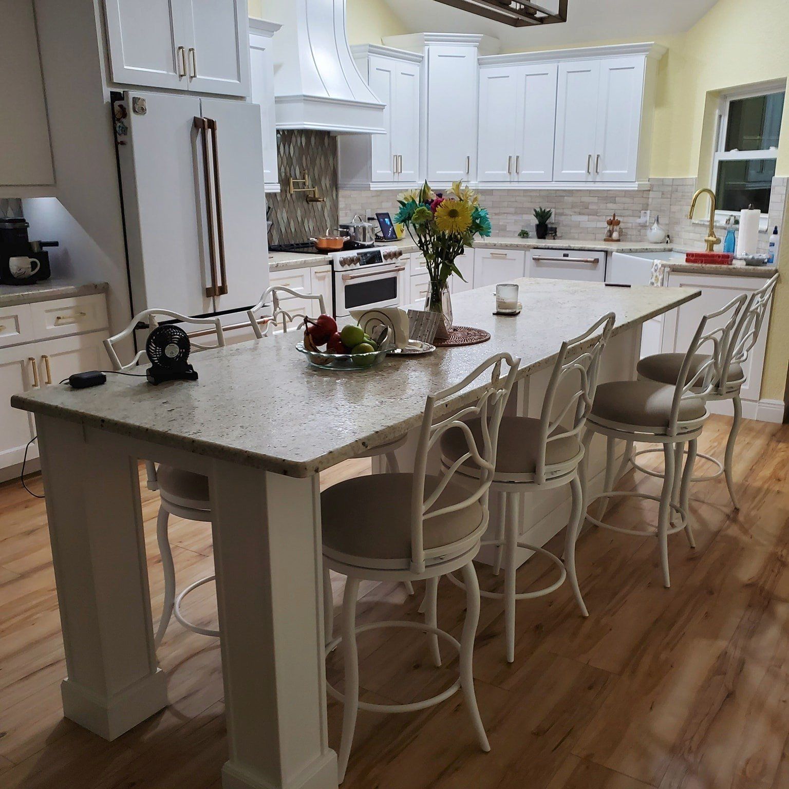 A bright kitchen features a large white island with light speckled countertops, four white bar stools, and wooden floors.