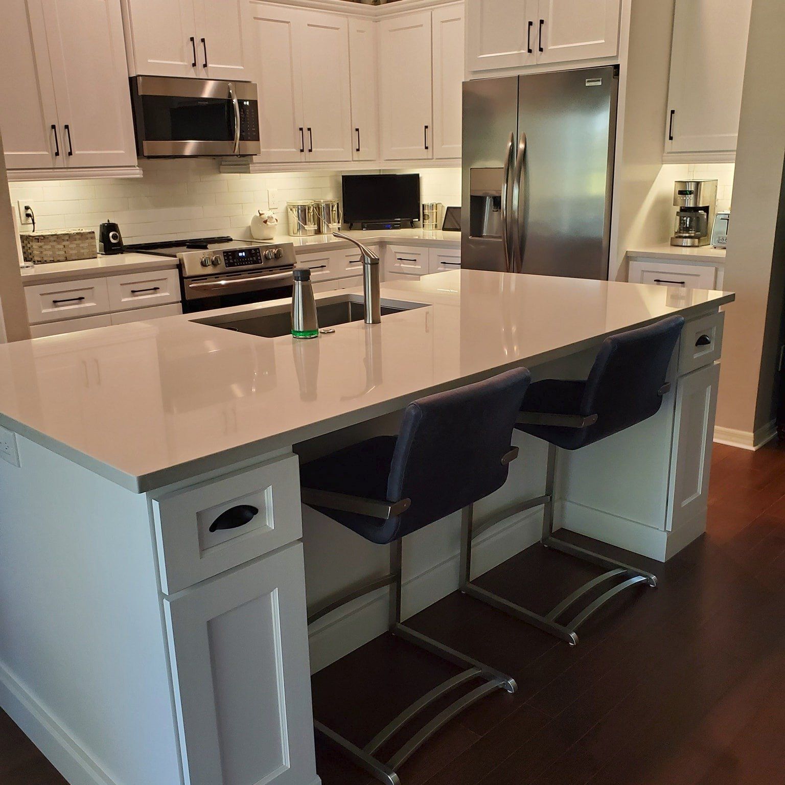 A modern kitchen with a white quartz island, two dark blue bar stools, white cabinetry, and stainless steel appliances.
