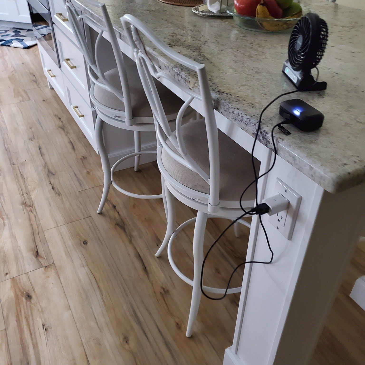 A small black desk fan sits on a kitchen island, plugged into an electrical outlet with a cord and a small adapter.