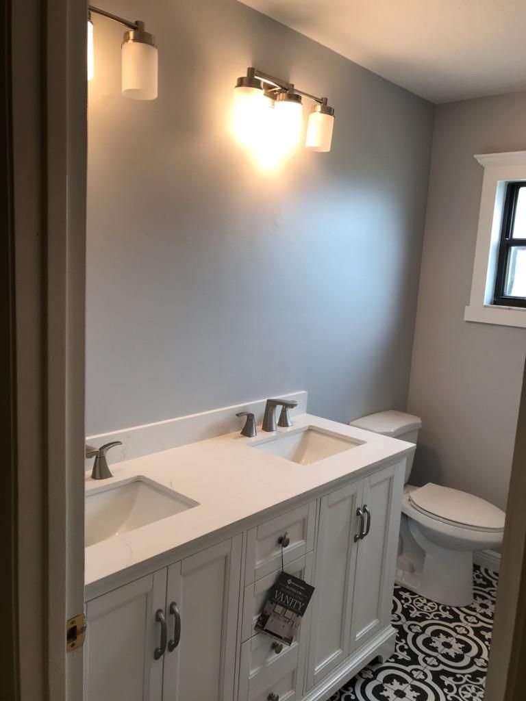 A bathroom with a white double-sink vanity, gray walls, dual light fixtures, and black-and-white patterned tile flooring.