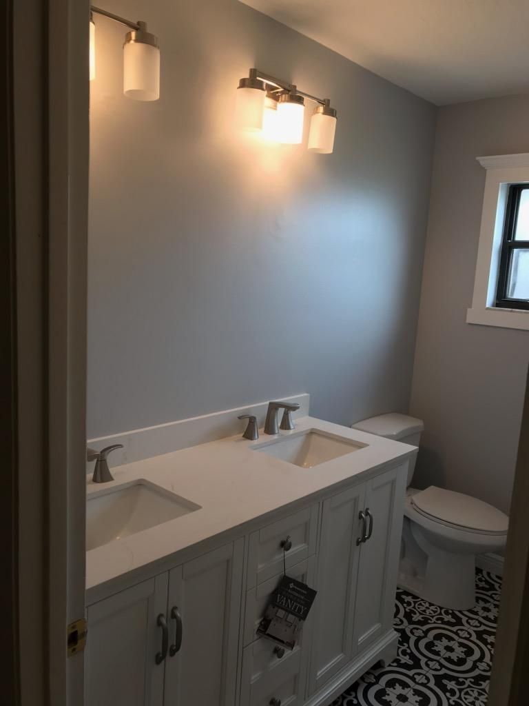 Bathroom with white double vanity, gray walls, two light fixtures, and a toilet on patterned black-and-white tile floor.