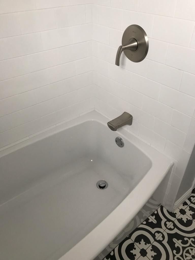 A white bathtub with brushed nickel hardware against white subway tile, featuring patterned black and white floor tiles.