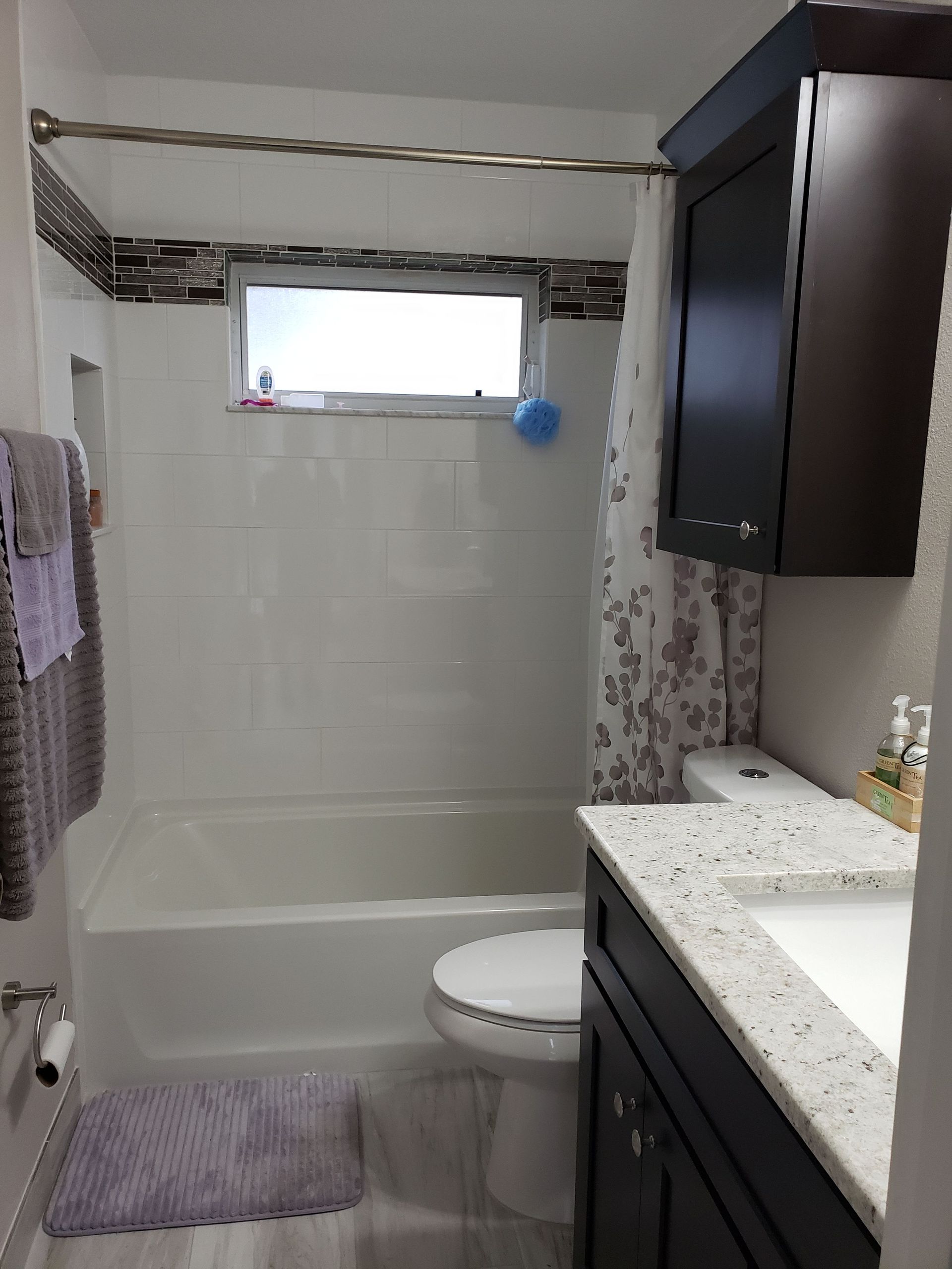 A small, modern bathroom featuring a white bathtub, toilet, and dark vanity with a speckled countertop and window.