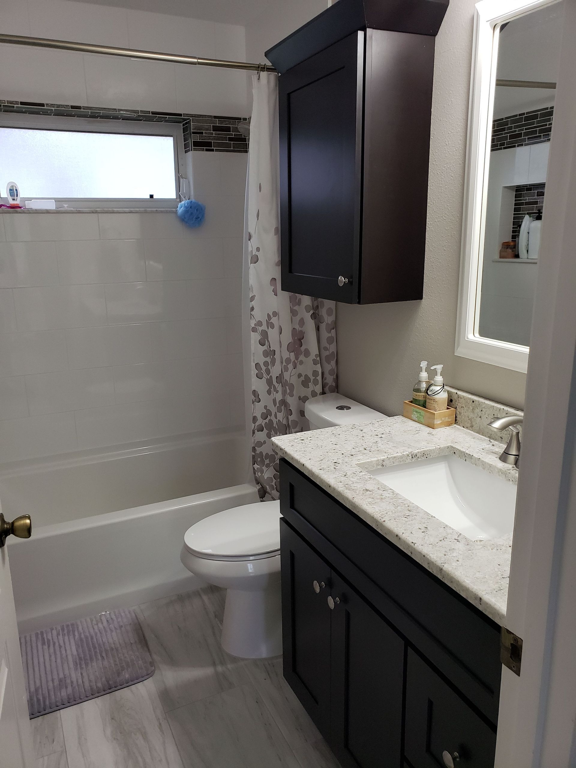 A small bathroom with dark wood cabinets, a speckled white vanity top, a toilet, and a bathtub with a floral shower curtain.