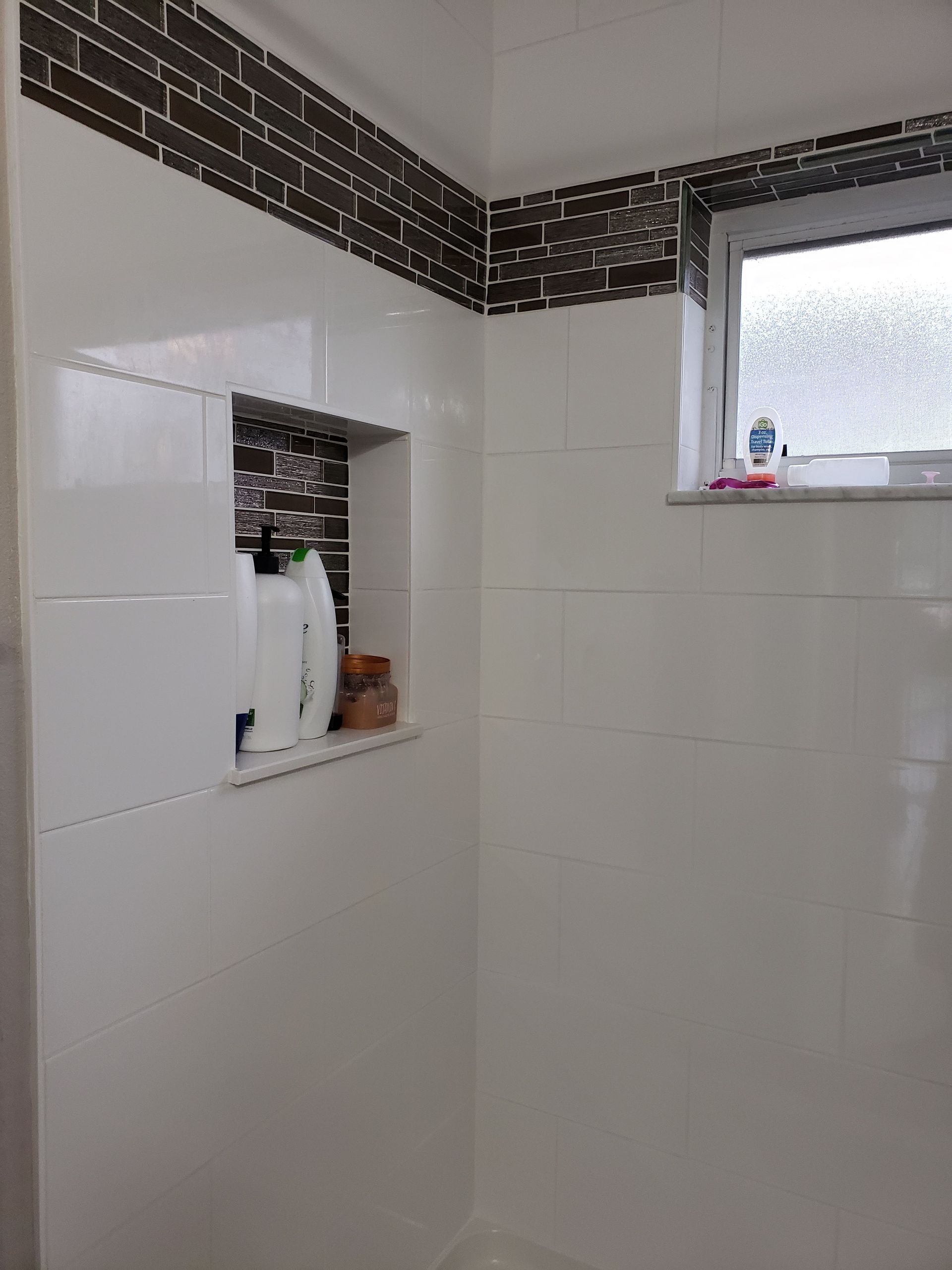 A shower corner with white tiled walls, a dark brown mosaic accent border, and a built-in recessed niche for toiletries.