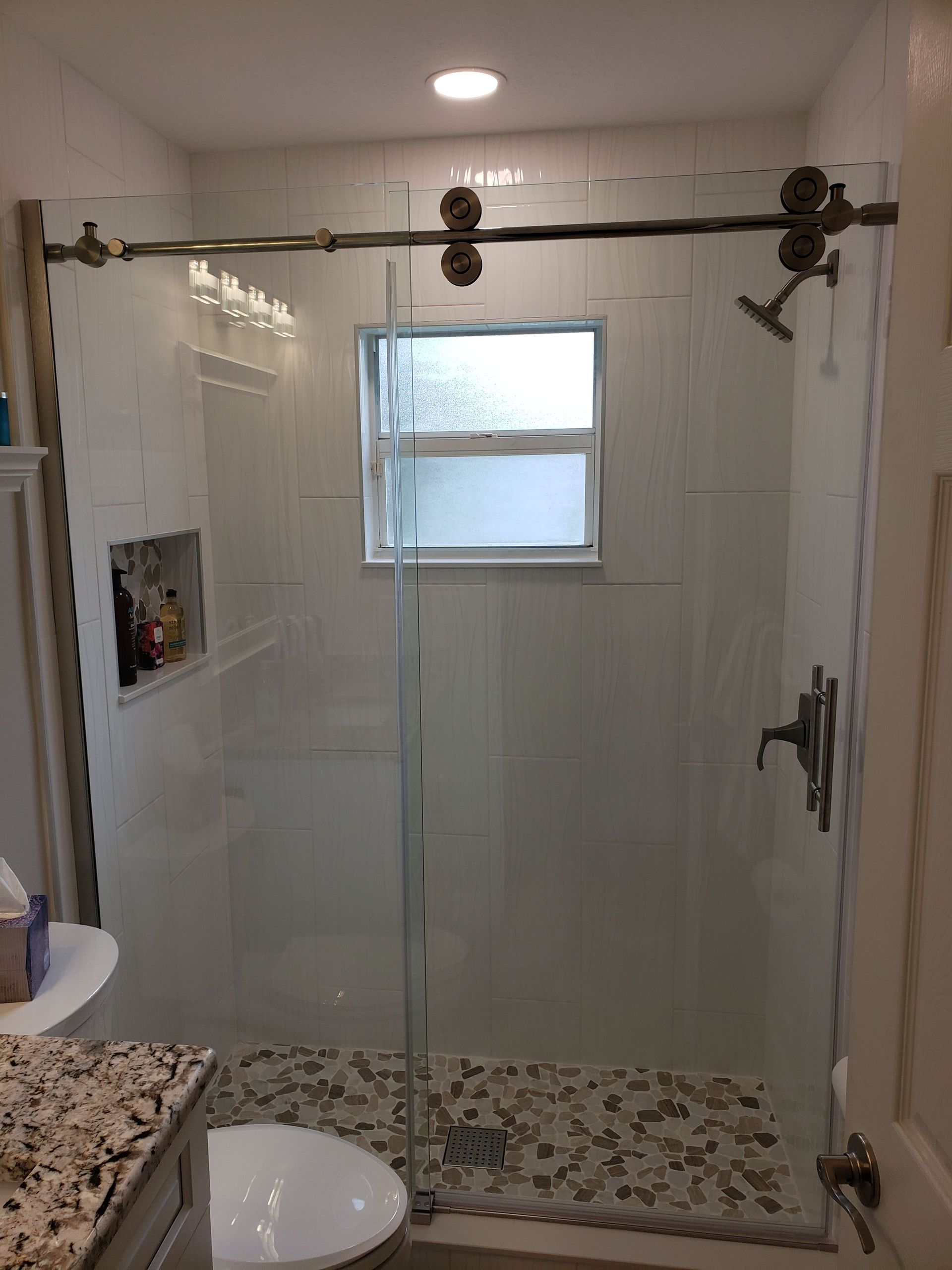 A modern walk-in shower with a glass sliding door, light tile walls, a pebble floor, and a built-in recessed shelf.