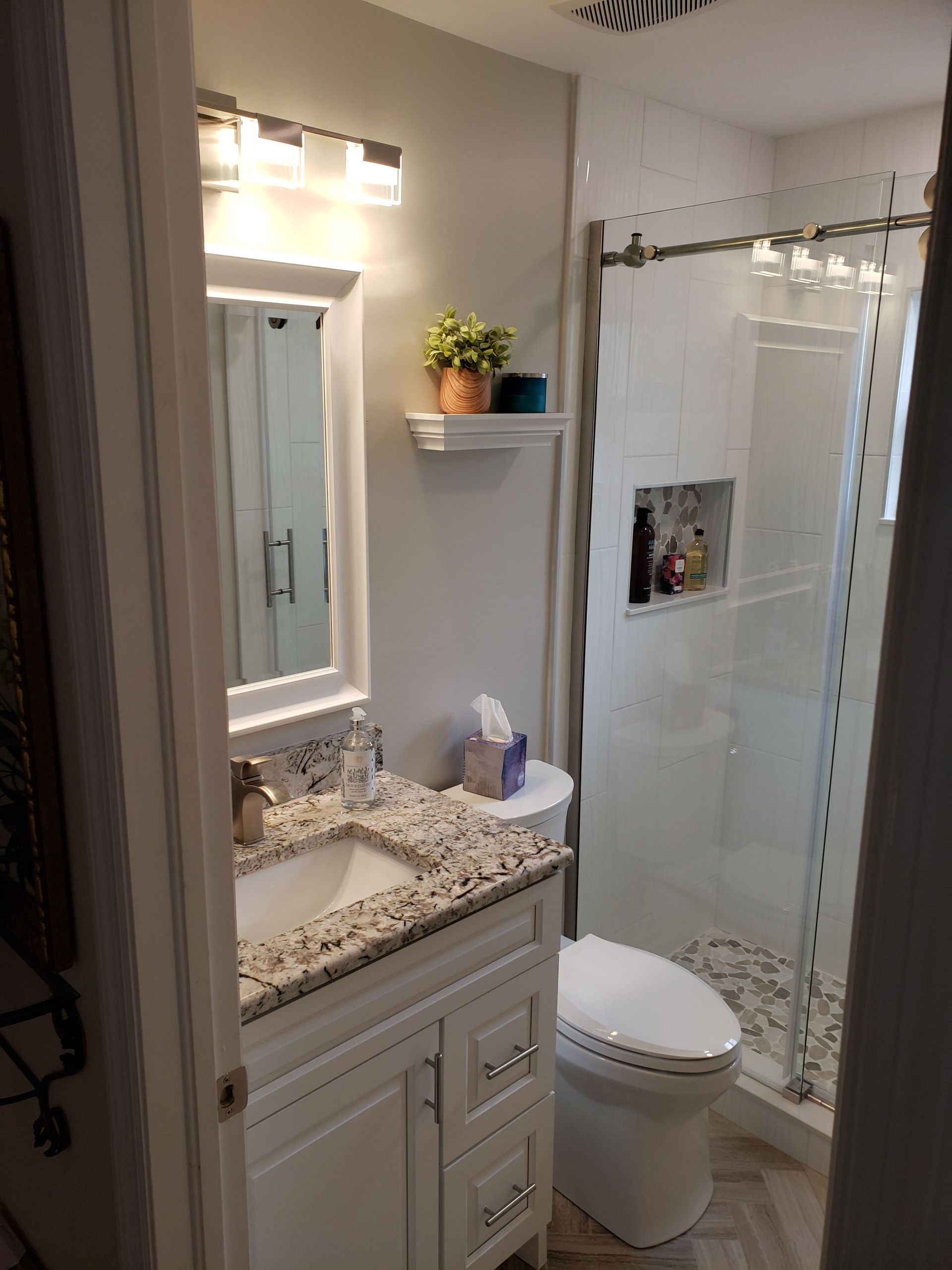 A compact bathroom featuring a white vanity with a granite countertop, a mirror, a toilet, and a glass-enclosed shower.