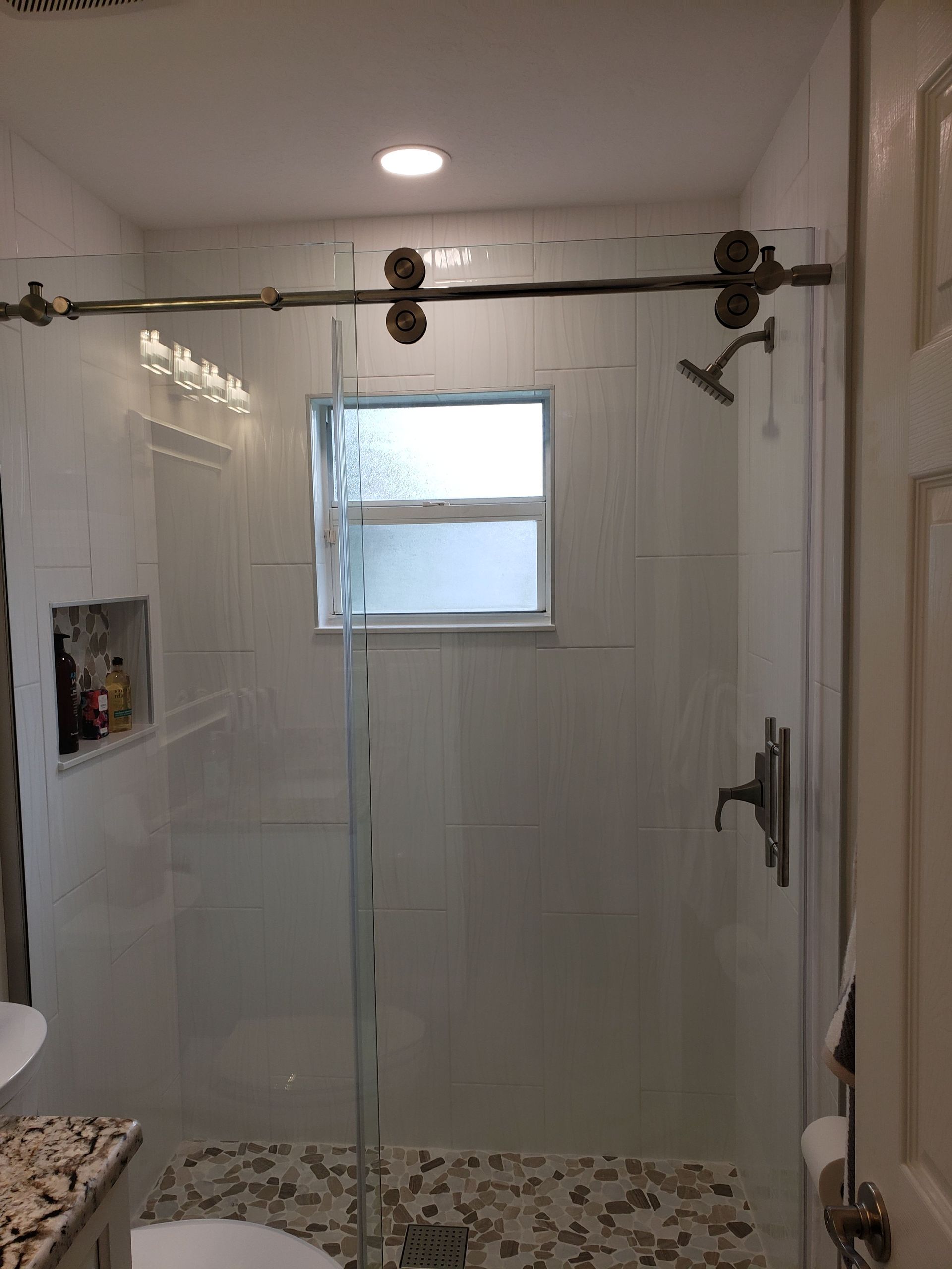 A modern walk-in shower with white tiled walls, a glass sliding door, a window, a pebble floor, and a metal shower head.