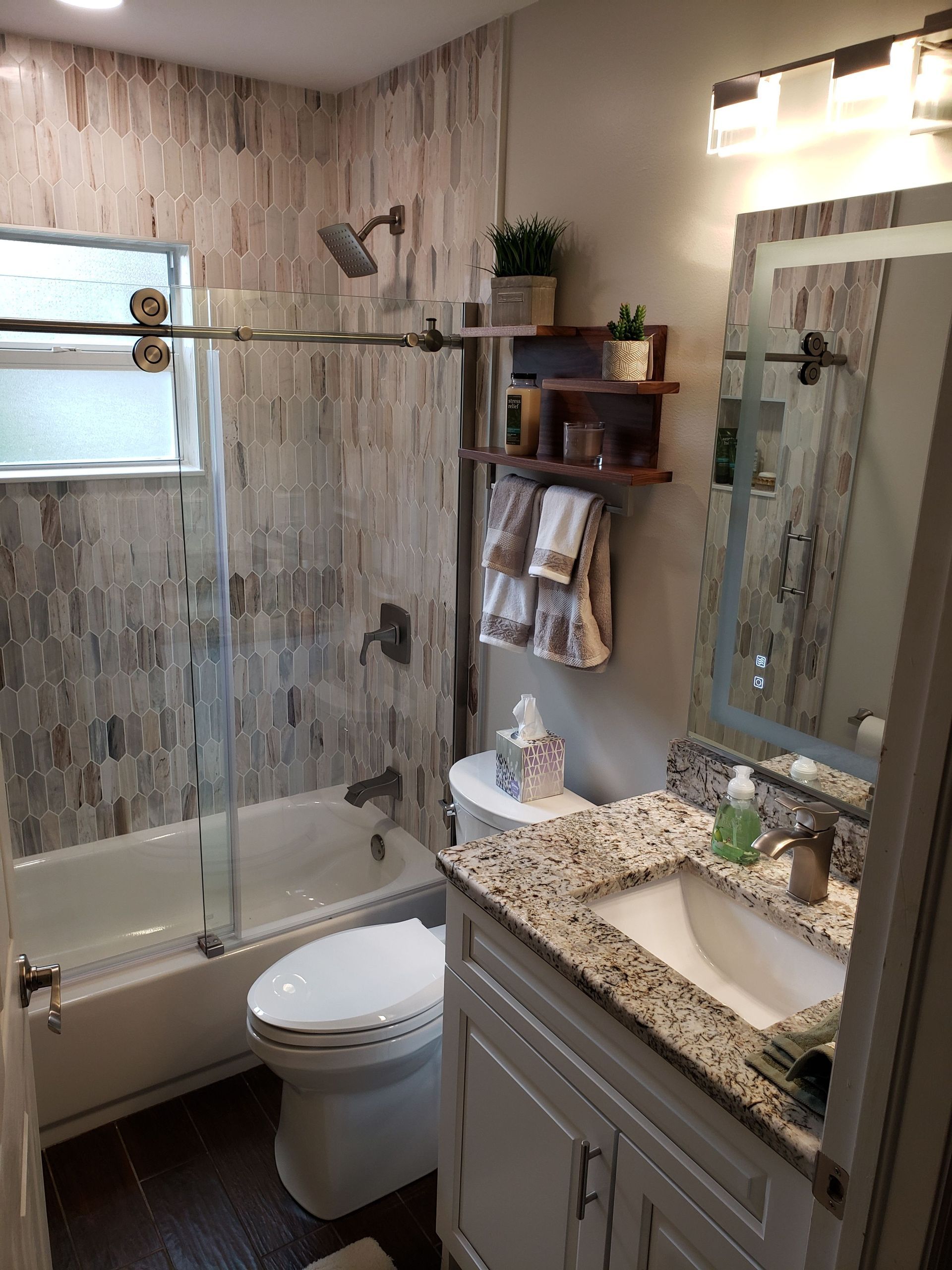 A bathroom with a granite-topped vanity, a white toilet, and a glass-enclosed shower with patterned tile walls.