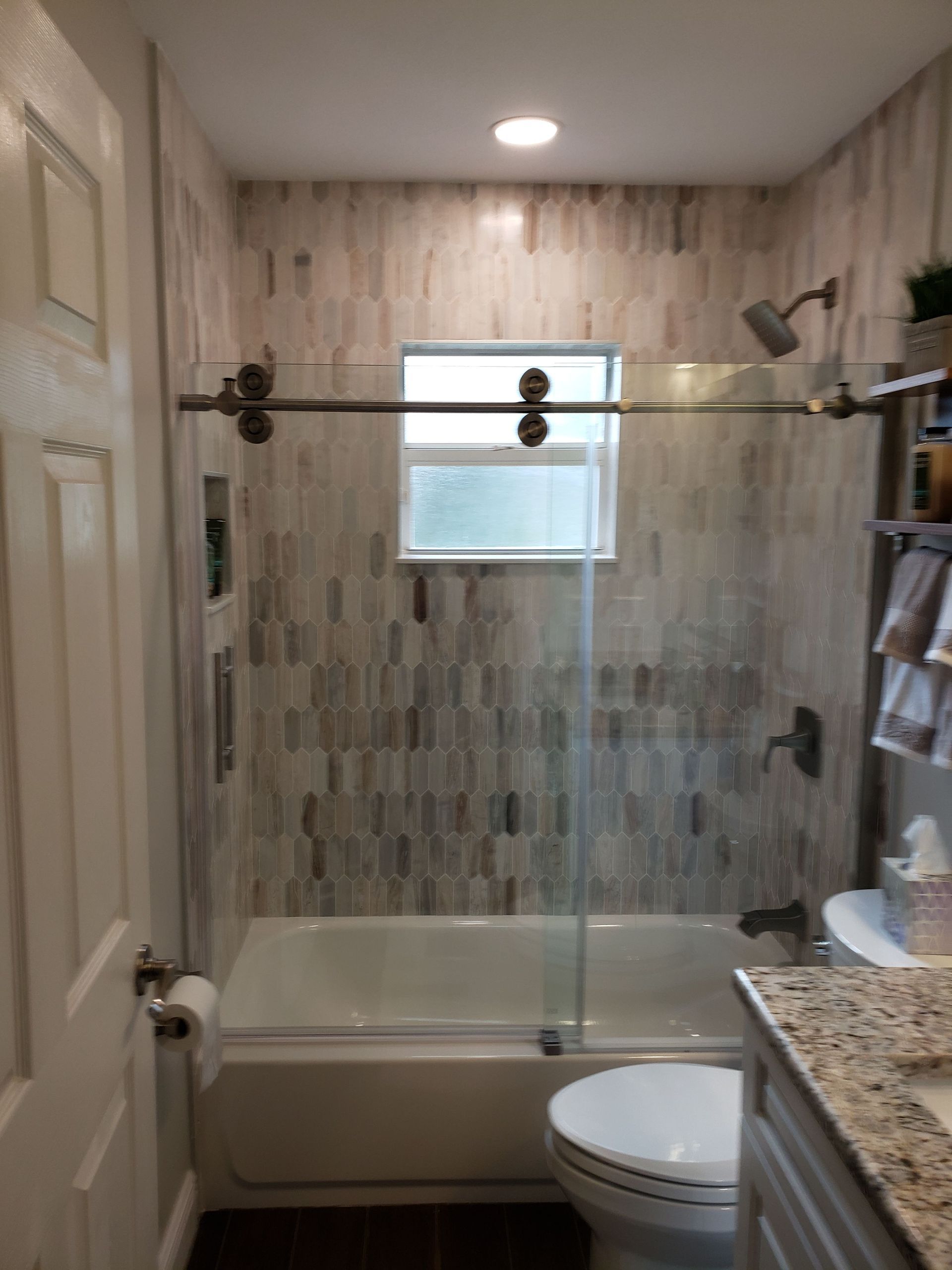 A bathroom with a bathtub featuring a glass sliding door, light-toned vertical patterned tile walls, and a window.