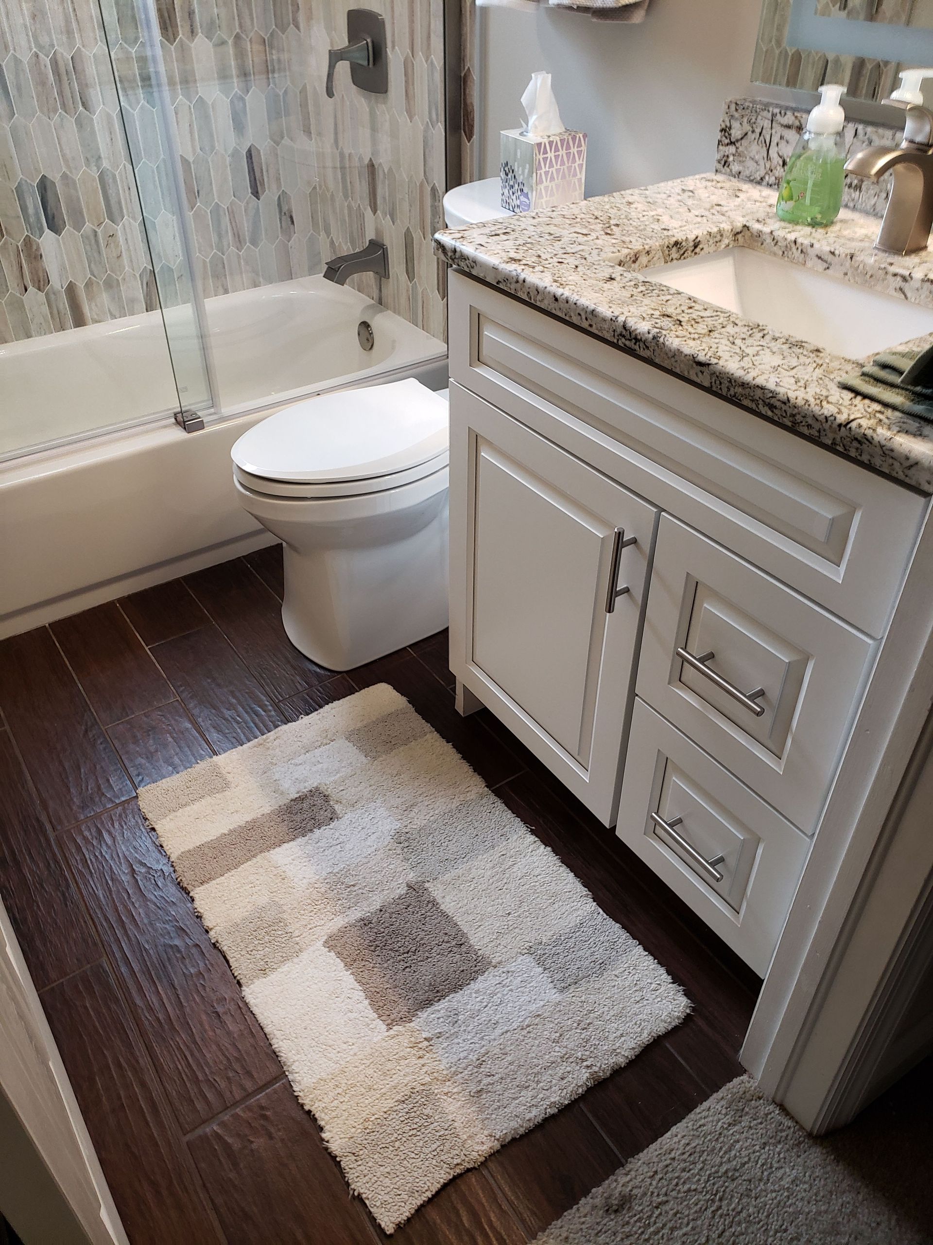 A modern bathroom with dark wood floors, a white vanity with a granite countertop, a toilet, and a bathtub with tile walls.
