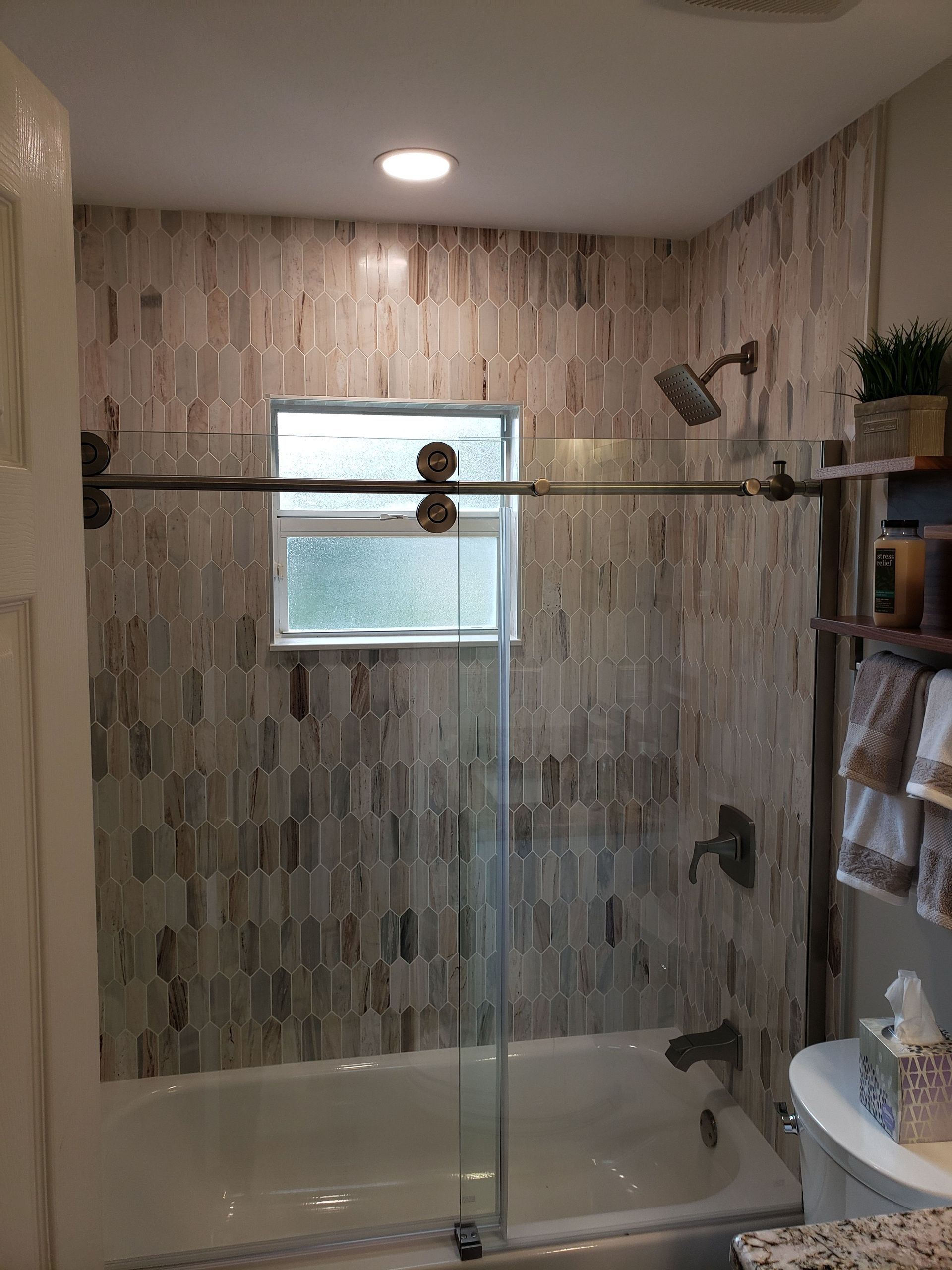 A bathroom with a bathtub, a sliding glass door, textured light-colored wall tiles, and a window above the tub.