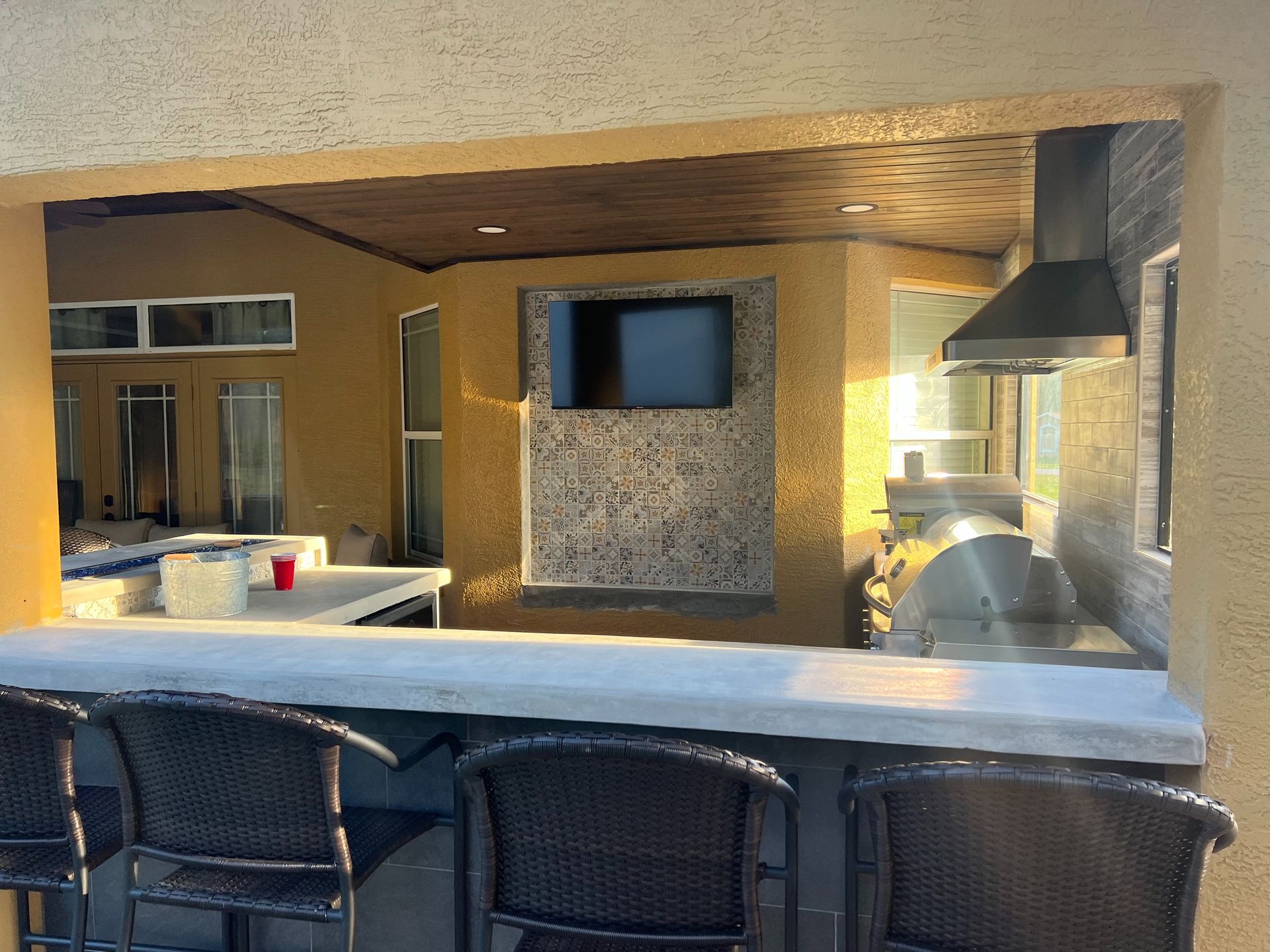Outdoor kitchen and bar area featuring a flat-screen TV mounted on a decorative tiled wall, seating, and a grill setup.