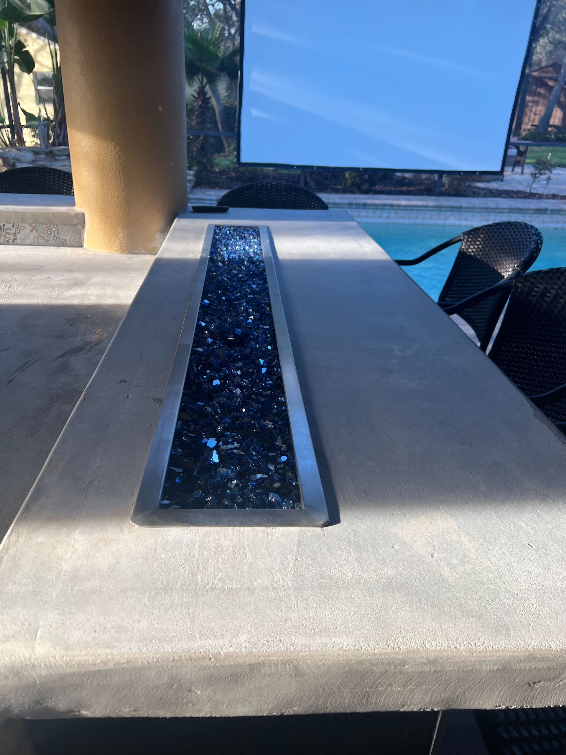 A linear fire pit filled with dark blue glass stones, set into a concrete bar top overlooking a pool and a large TV.