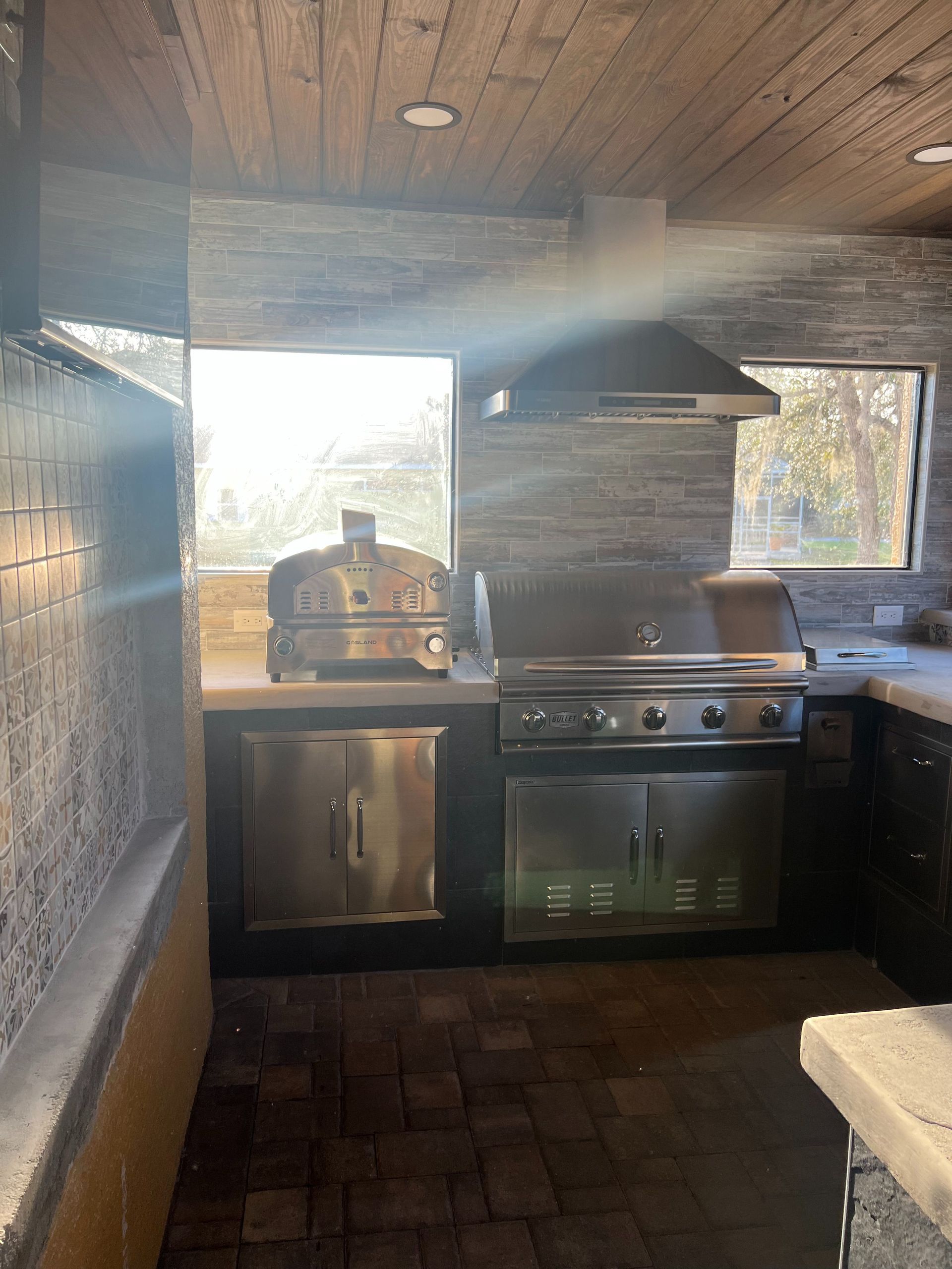 Outdoor kitchen with a stainless steel grill and pizza oven, stone walls, and a wood ceiling.