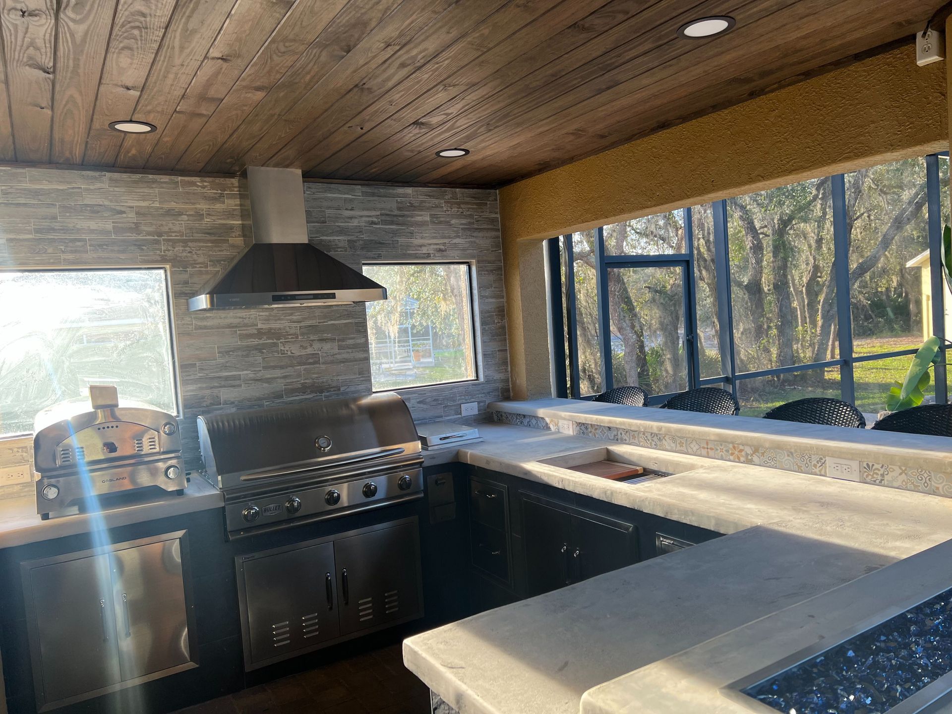 An outdoor kitchen with a stainless steel grill, vent hood, stone backsplash, and concrete countertops under a wood ceiling.