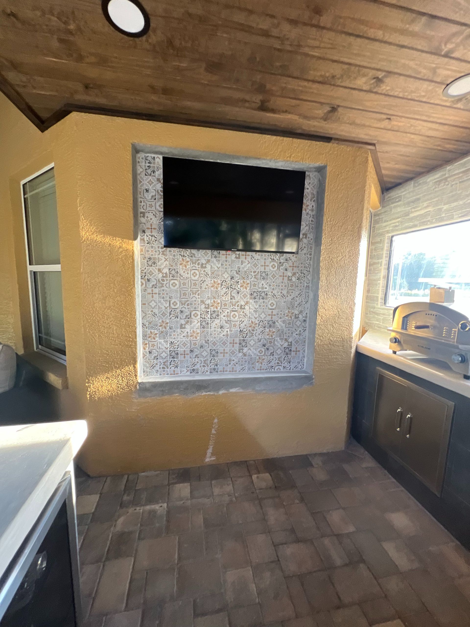 A covered outdoor patio featuring a mounted television, decorative tiled wall, pizza oven, and paver flooring.