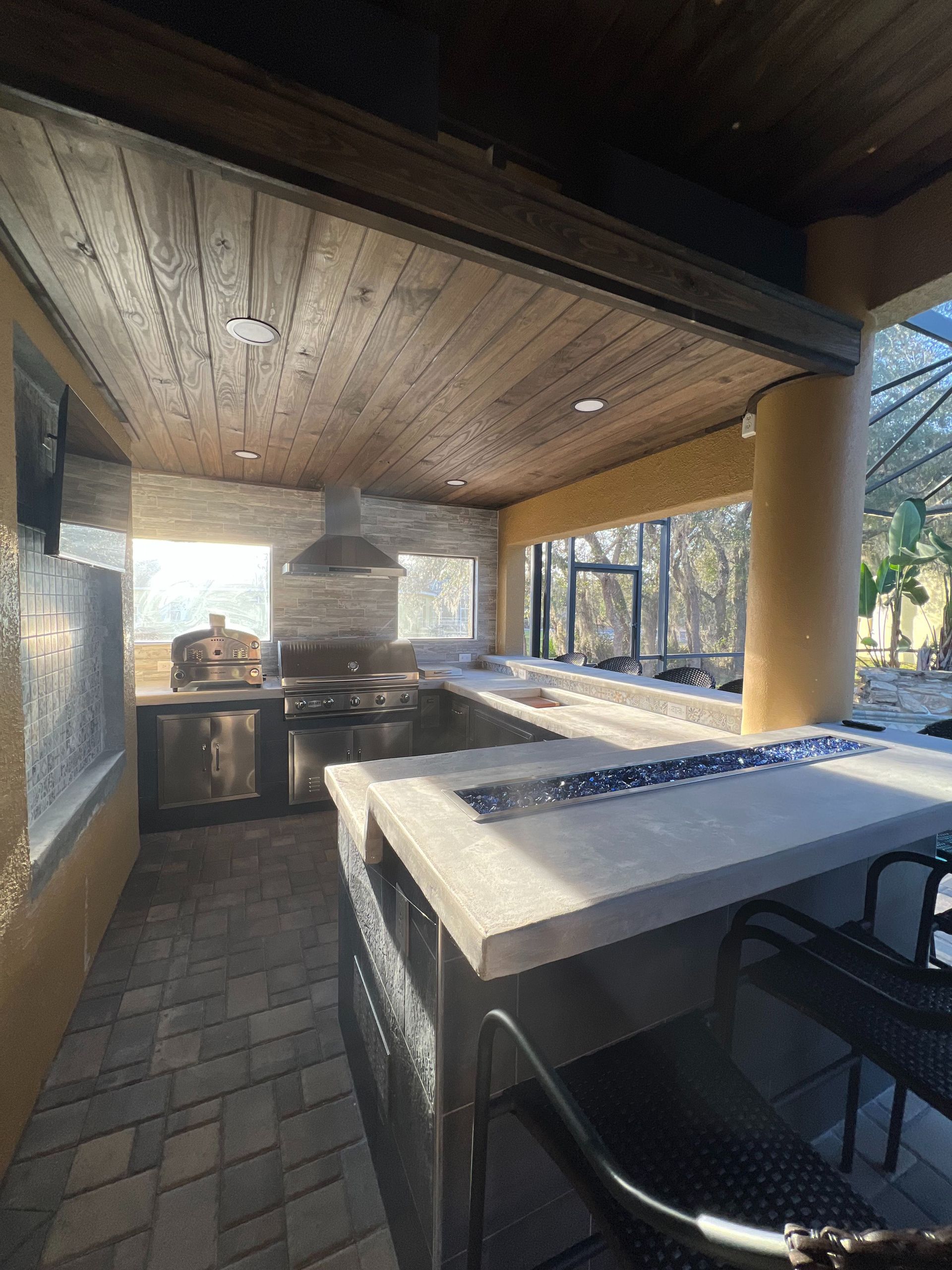 An outdoor covered kitchen features a stone island, pizza oven, built-in grill, and wood-paneled ceiling.