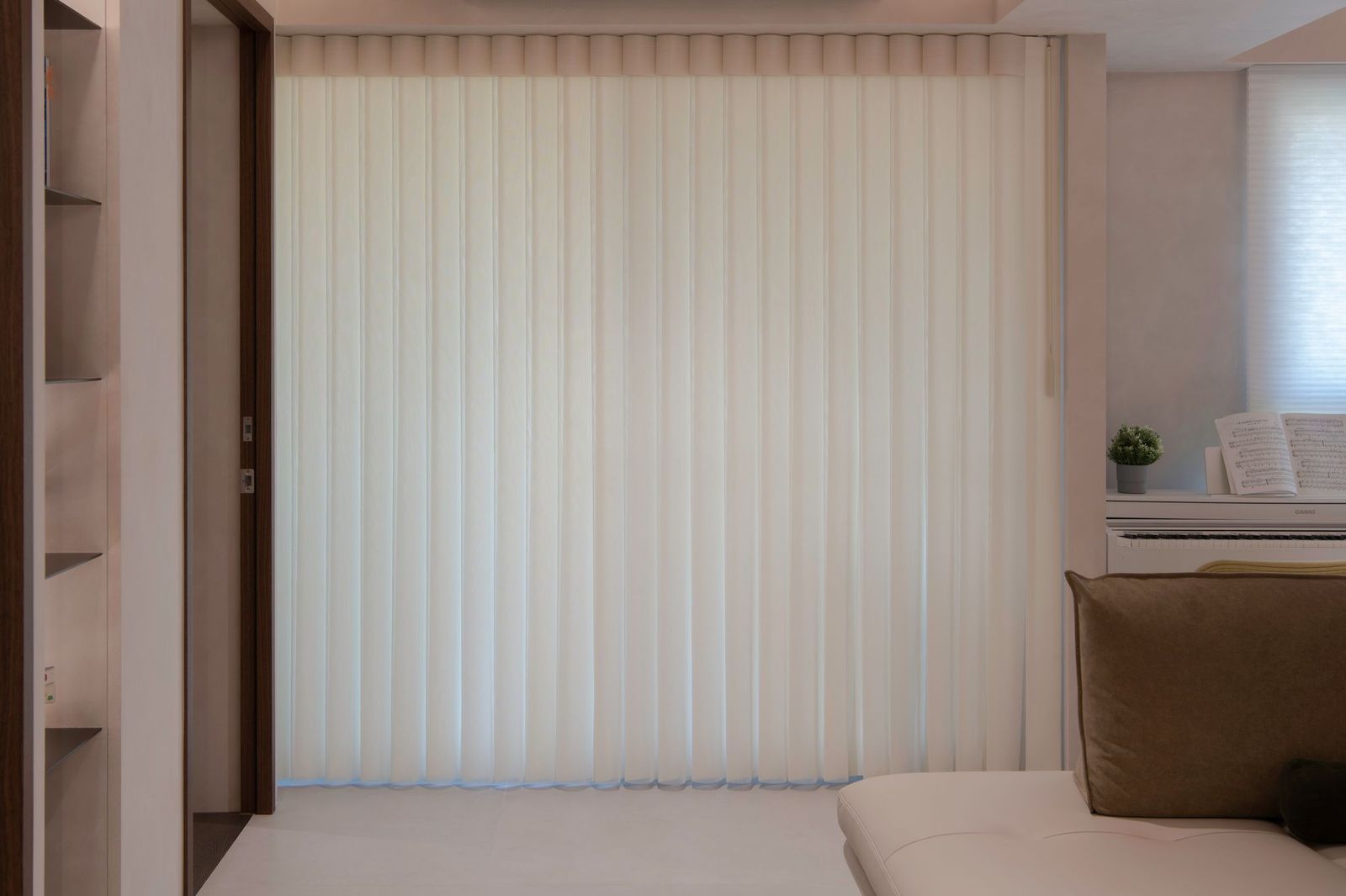 Floor-to-ceiling white vertical blinds cover a large window in a modern, neutral-toned living room.