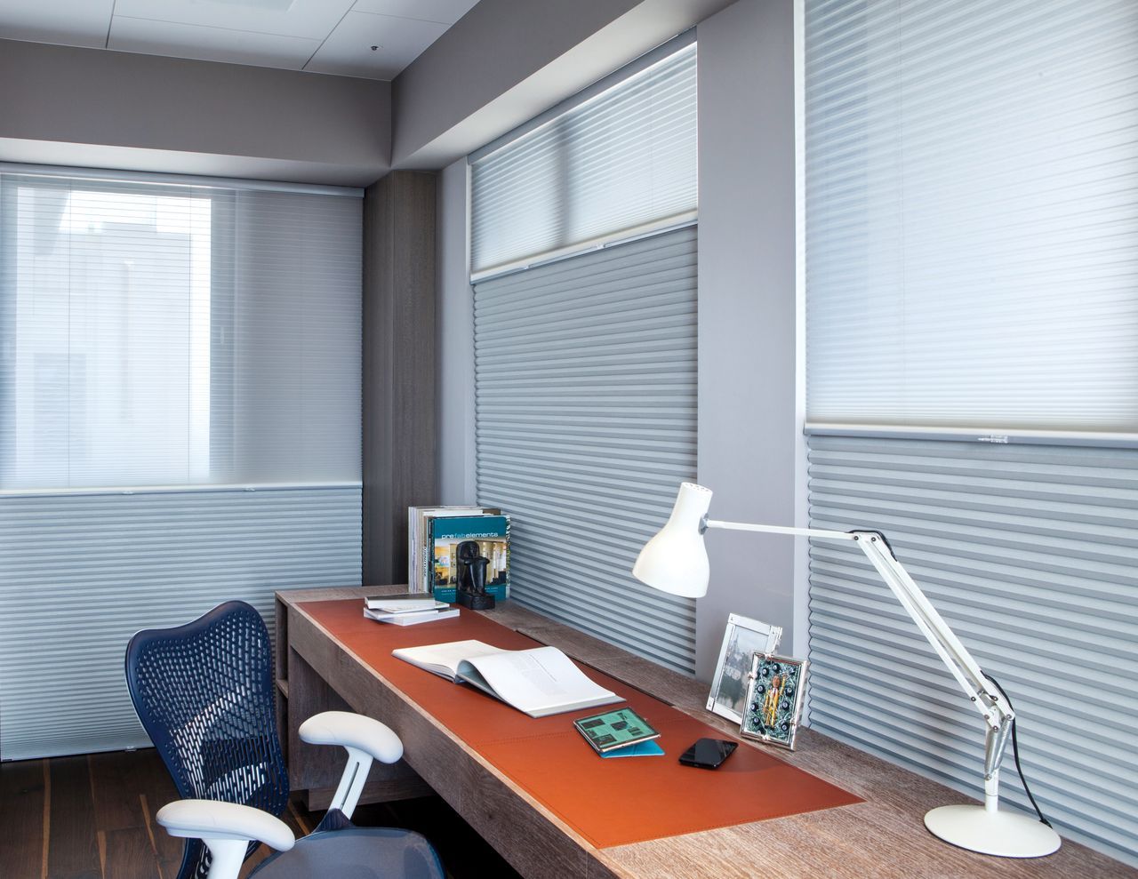 A home office with a long, brown desk, white task lamp, blue office chair, and windows covered by pleated blinds.
