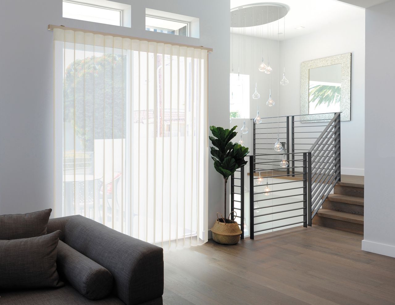 A modern living room with a grey sofa, tall white vertical blinds on a sliding door, a potted plant, and a staircase.