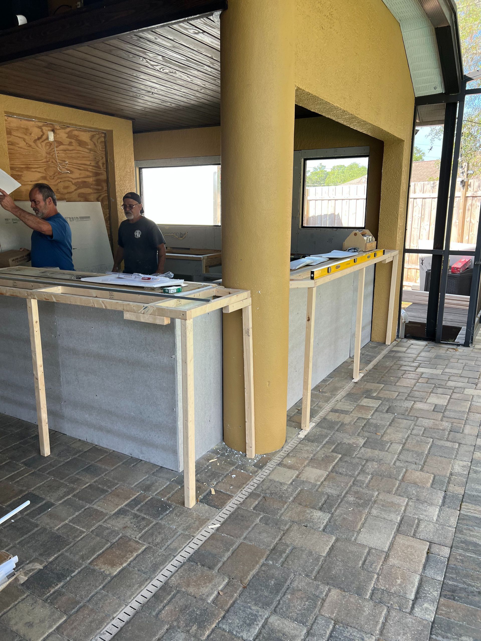 Two workers construct a stone-clad outdoor bar frame with yellow stucco columns under a covered patio.