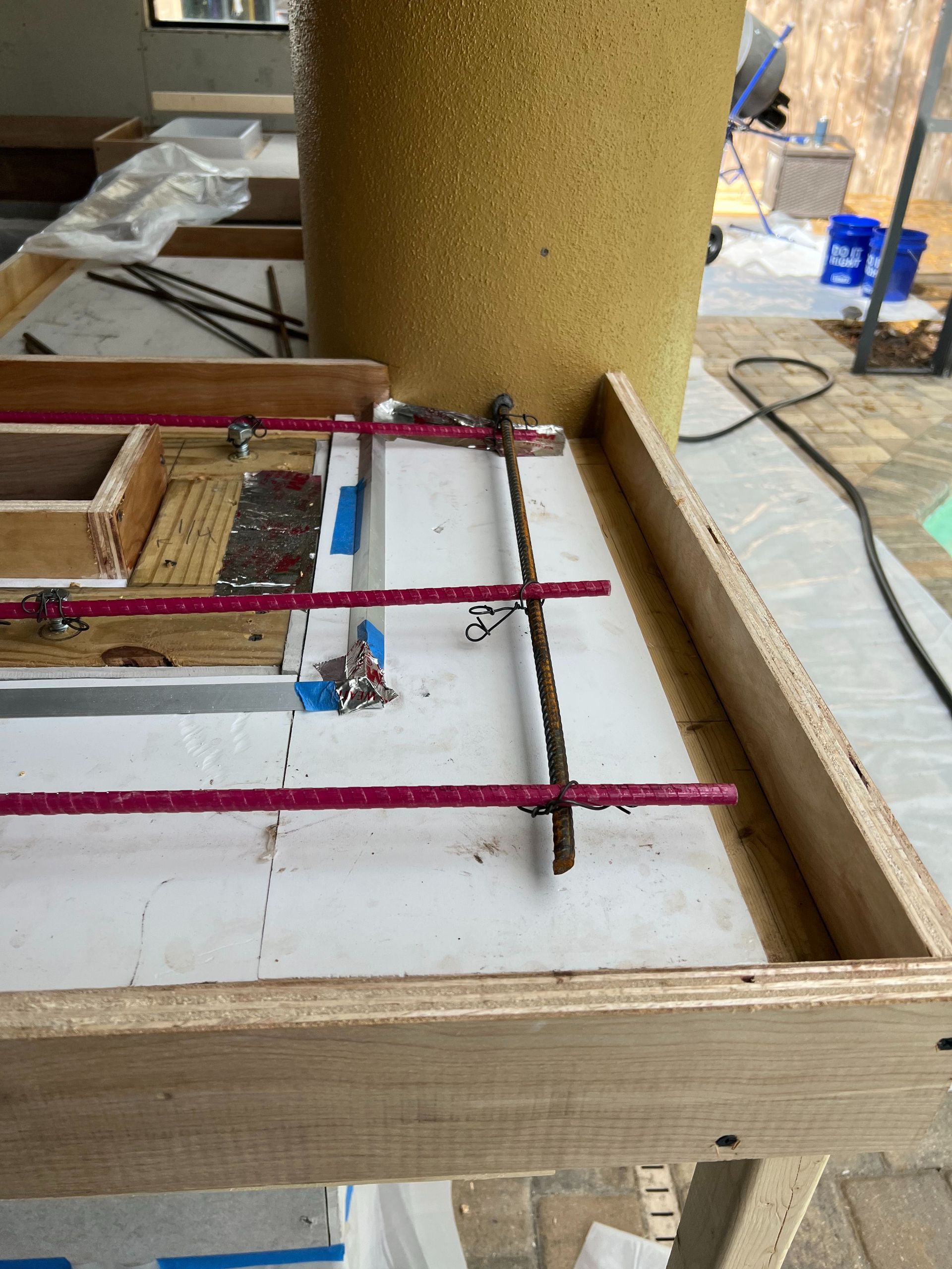 Wooden concrete formwork with red rebar set up on a white surface, with a large tan cylindrical form in the background.