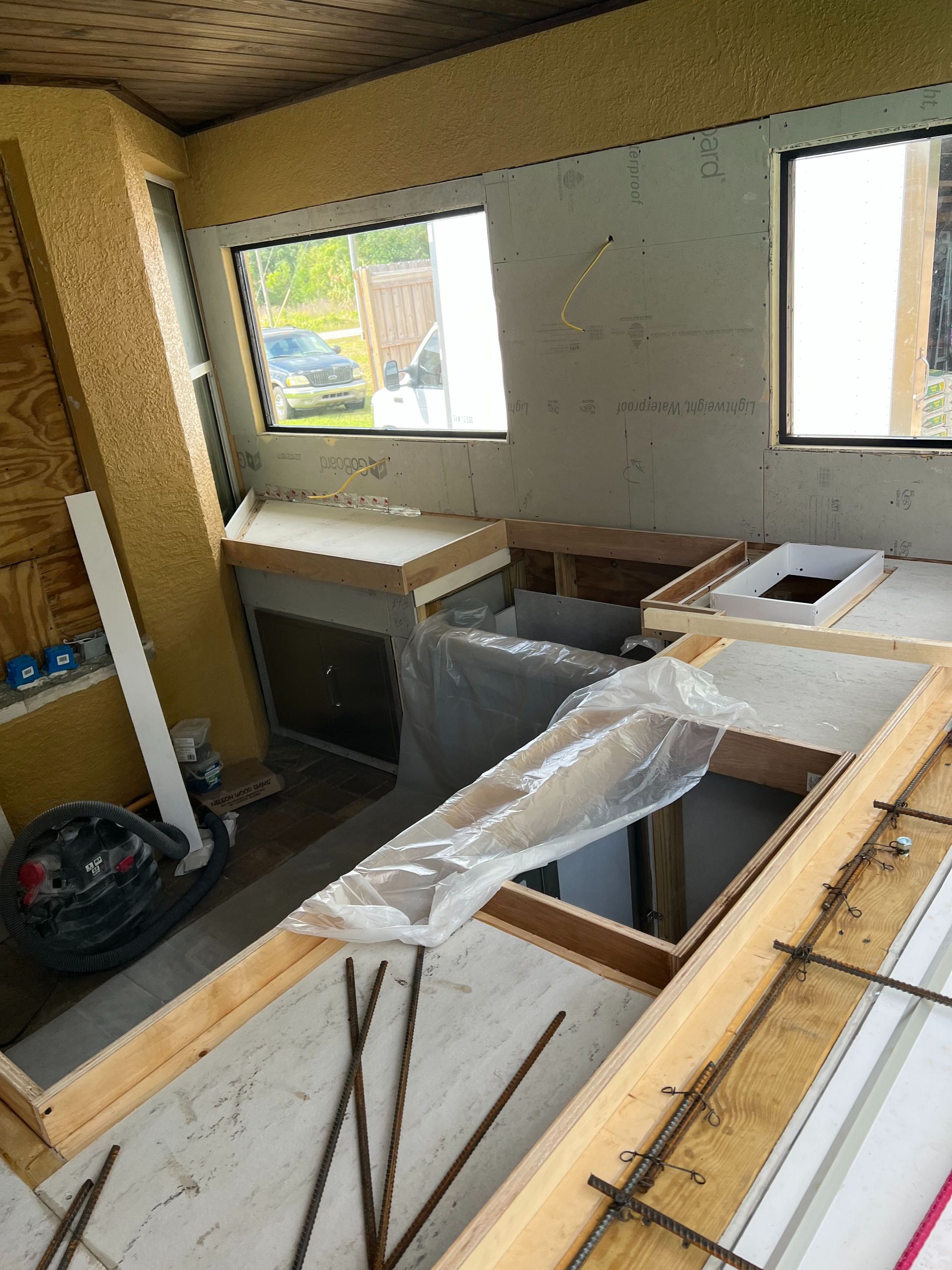 An unfinished kitchen renovation showing wooden frames for countertops, a plastic-covered sink area, and two windows.