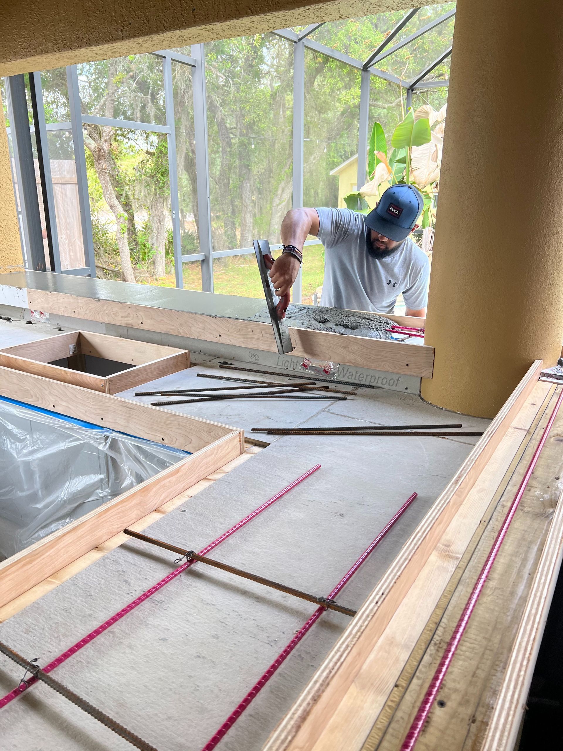 A person in a gray shirt and blue cap uses a tool to secure metal rebar inside a wooden form for a concrete project.