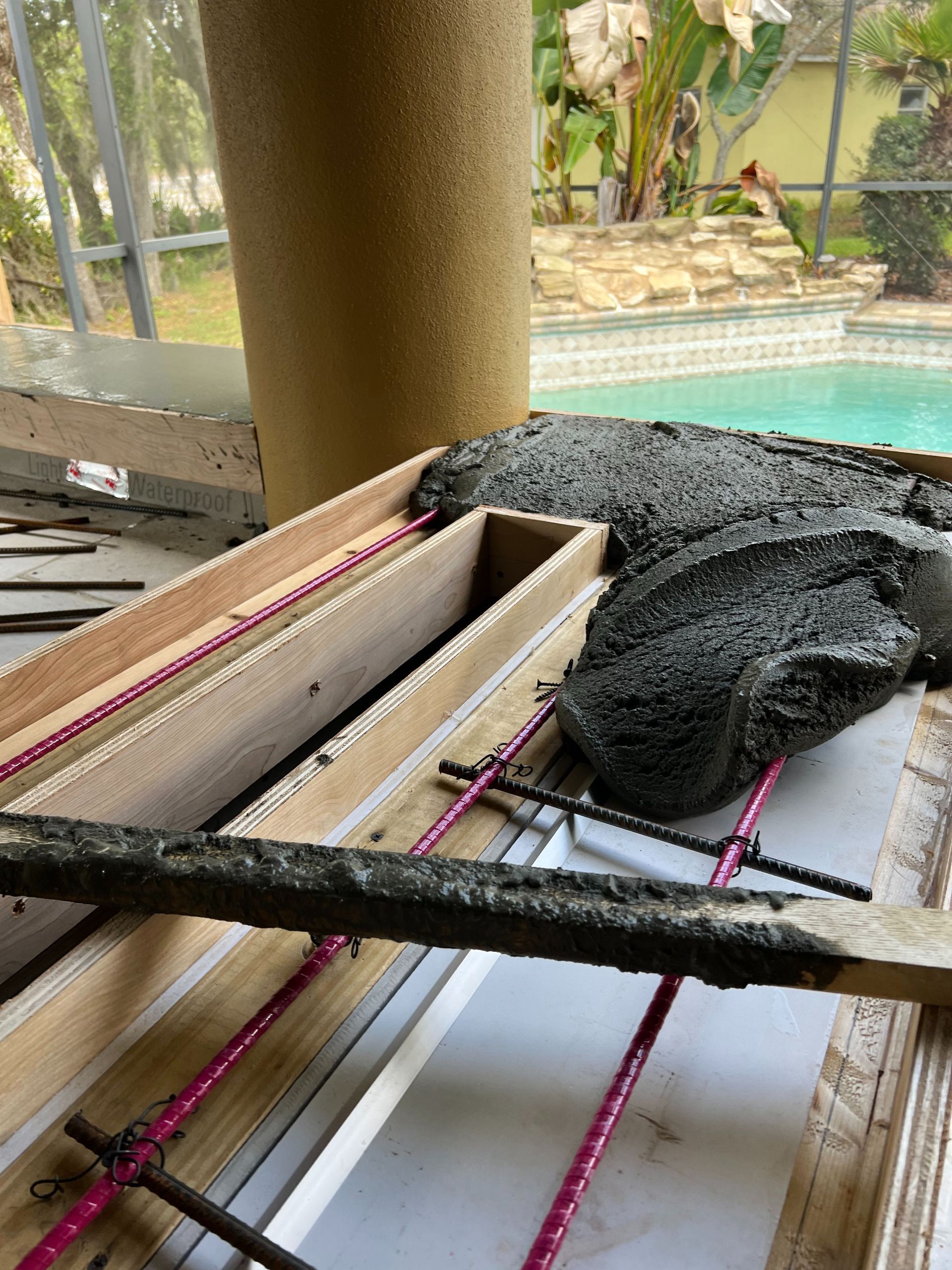 Wet concrete being poured into a wooden form containing pink fiberglass rebar, with a swimming pool in the background.