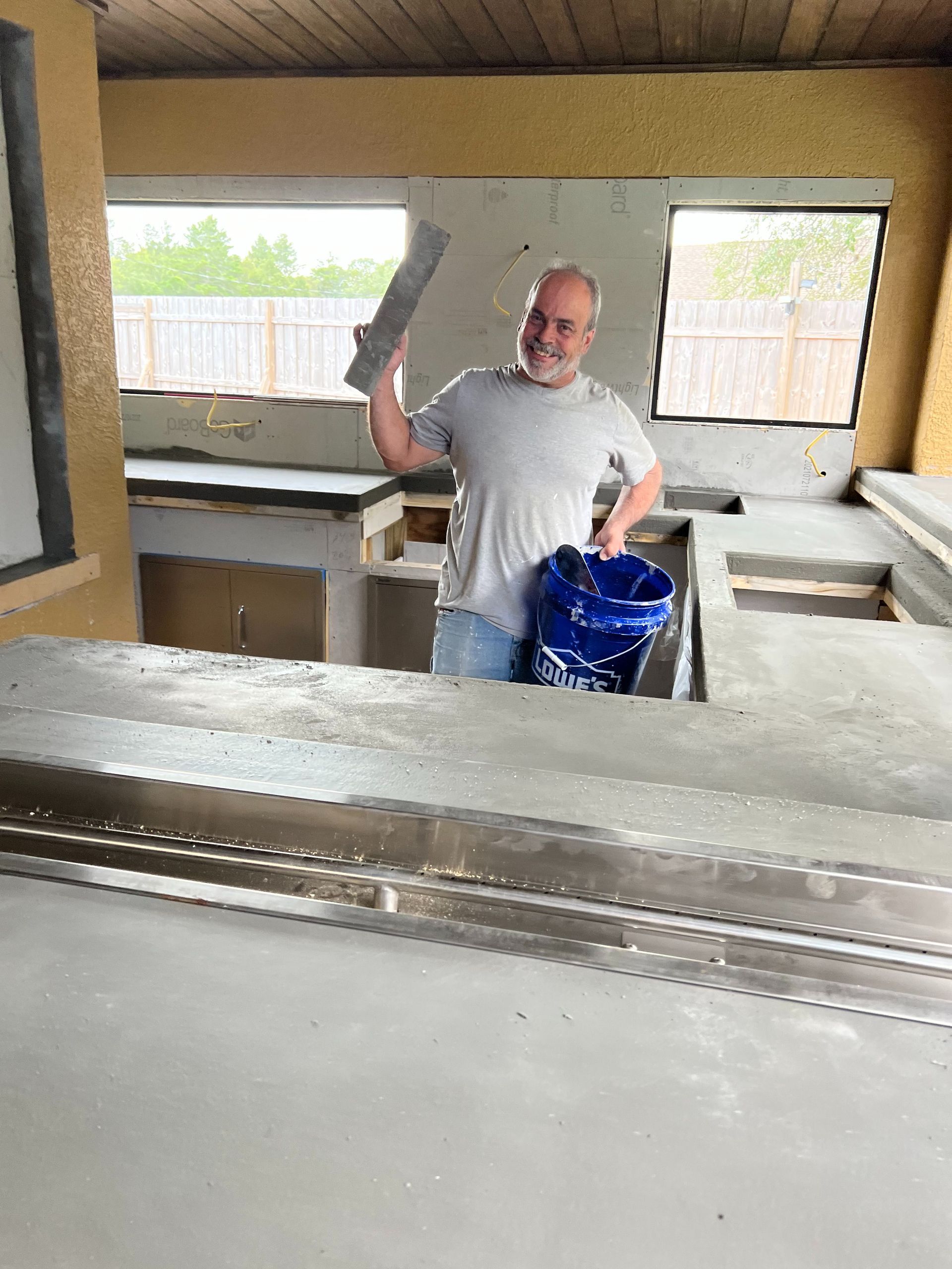 A person holding a tool and a blue bucket stands in a room under construction with concrete counters and window openings.