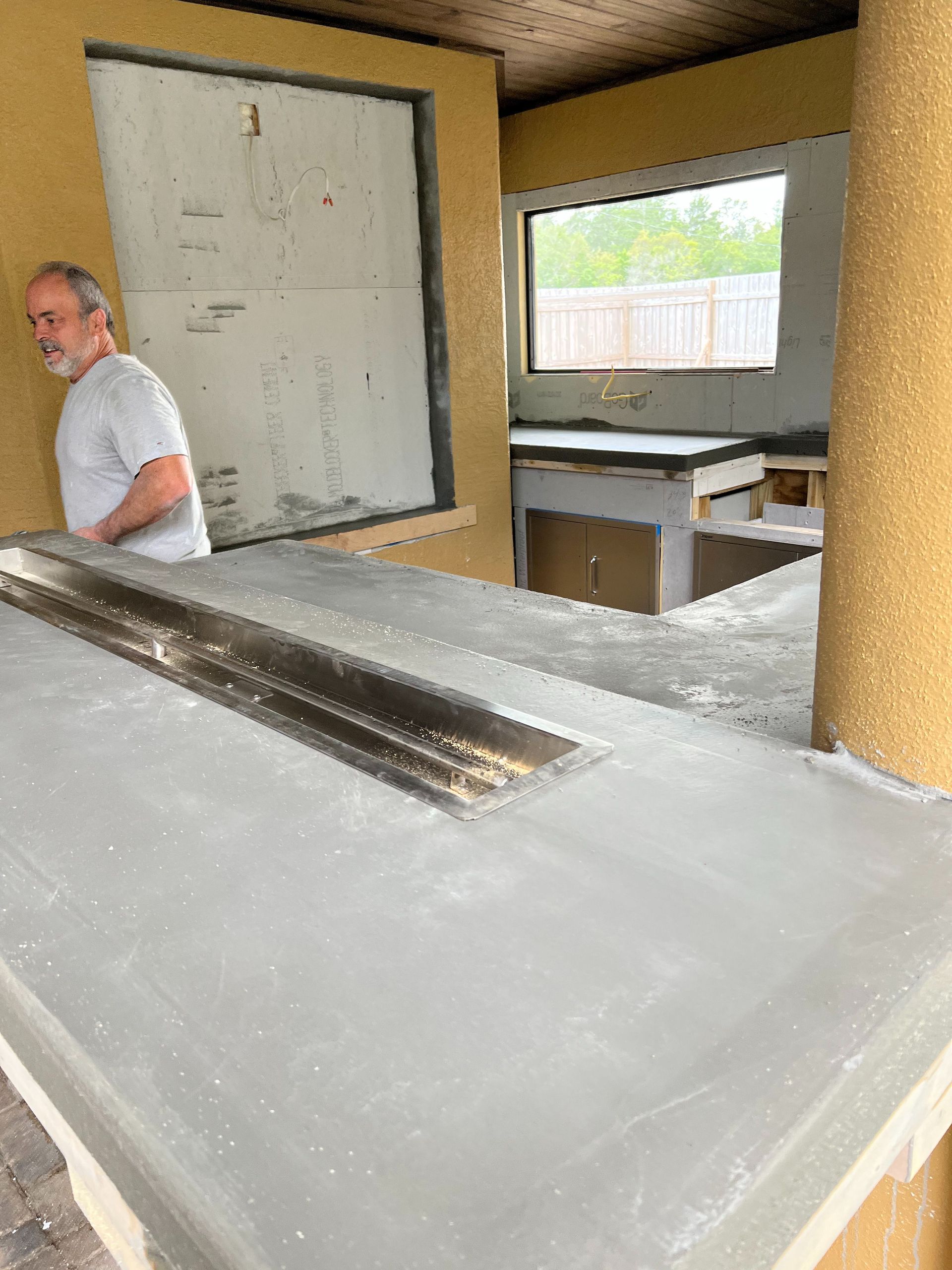 A person stands near a gray concrete countertop with a recessed rectangular metal trough in an outdoor kitchen setting.