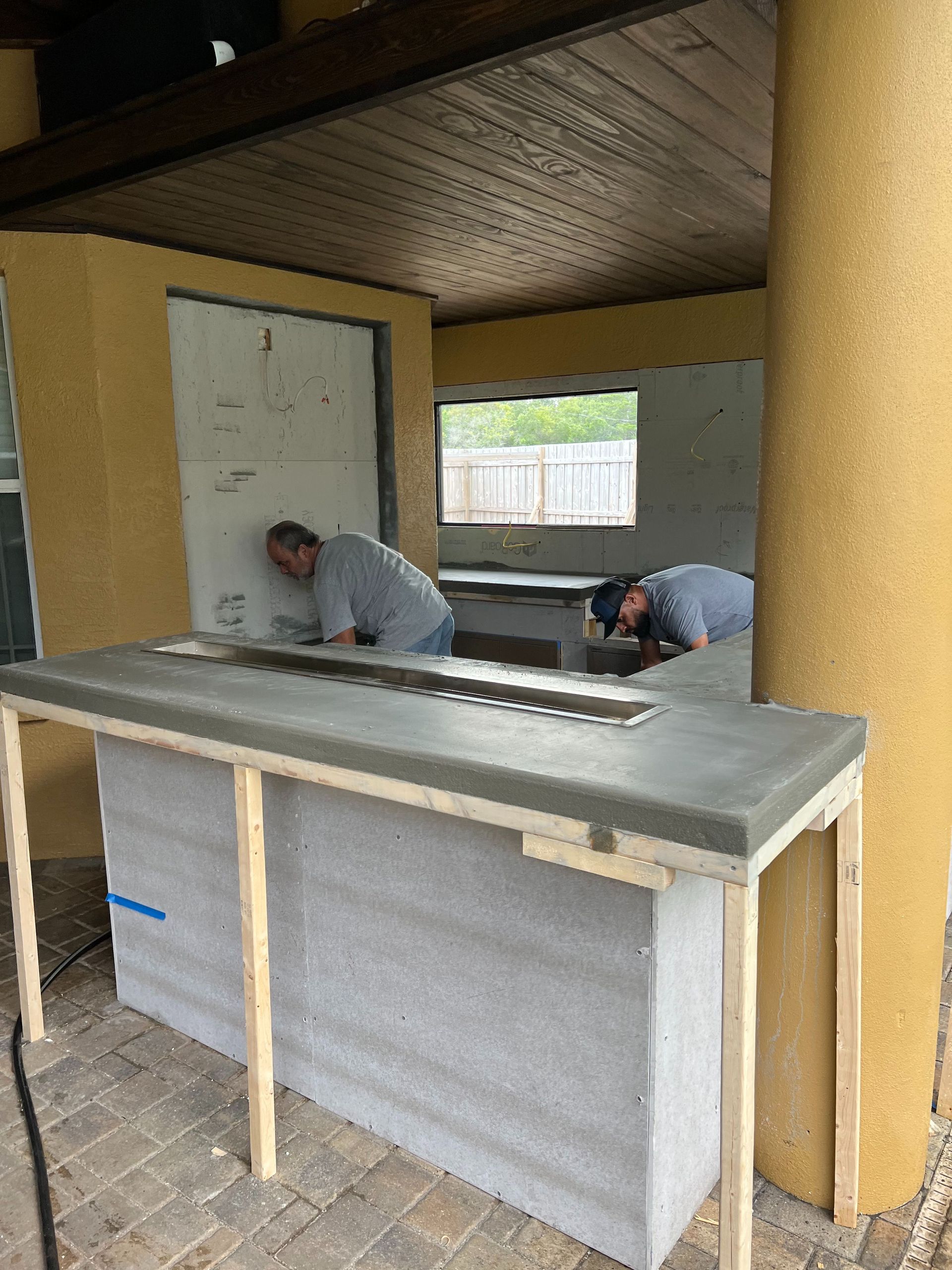 Two workers building a concrete outdoor kitchen bar under a covered patio.