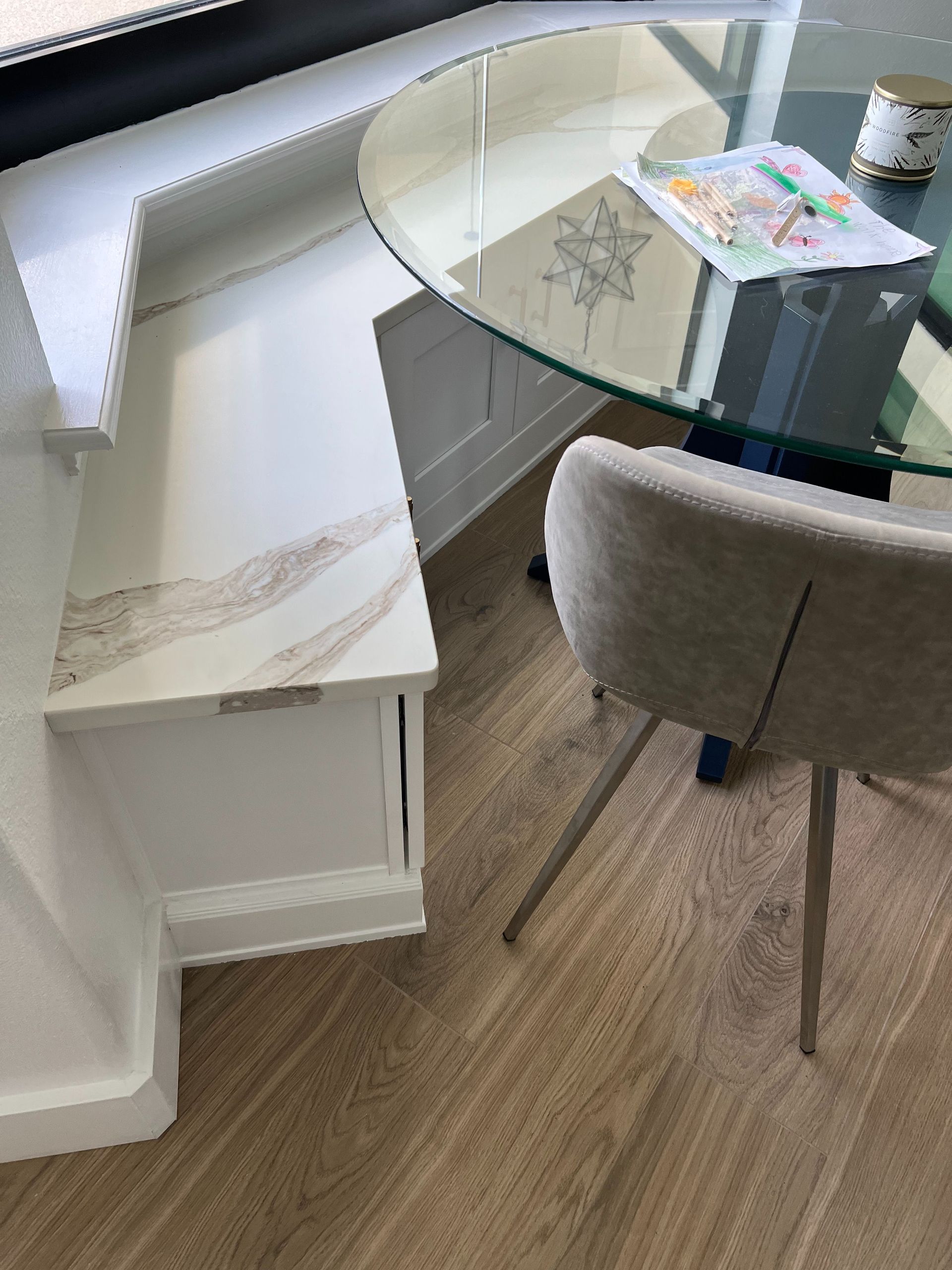 A white built-in corner bench with a marble top next to a glass dining table and a grey chair on a wood floor.