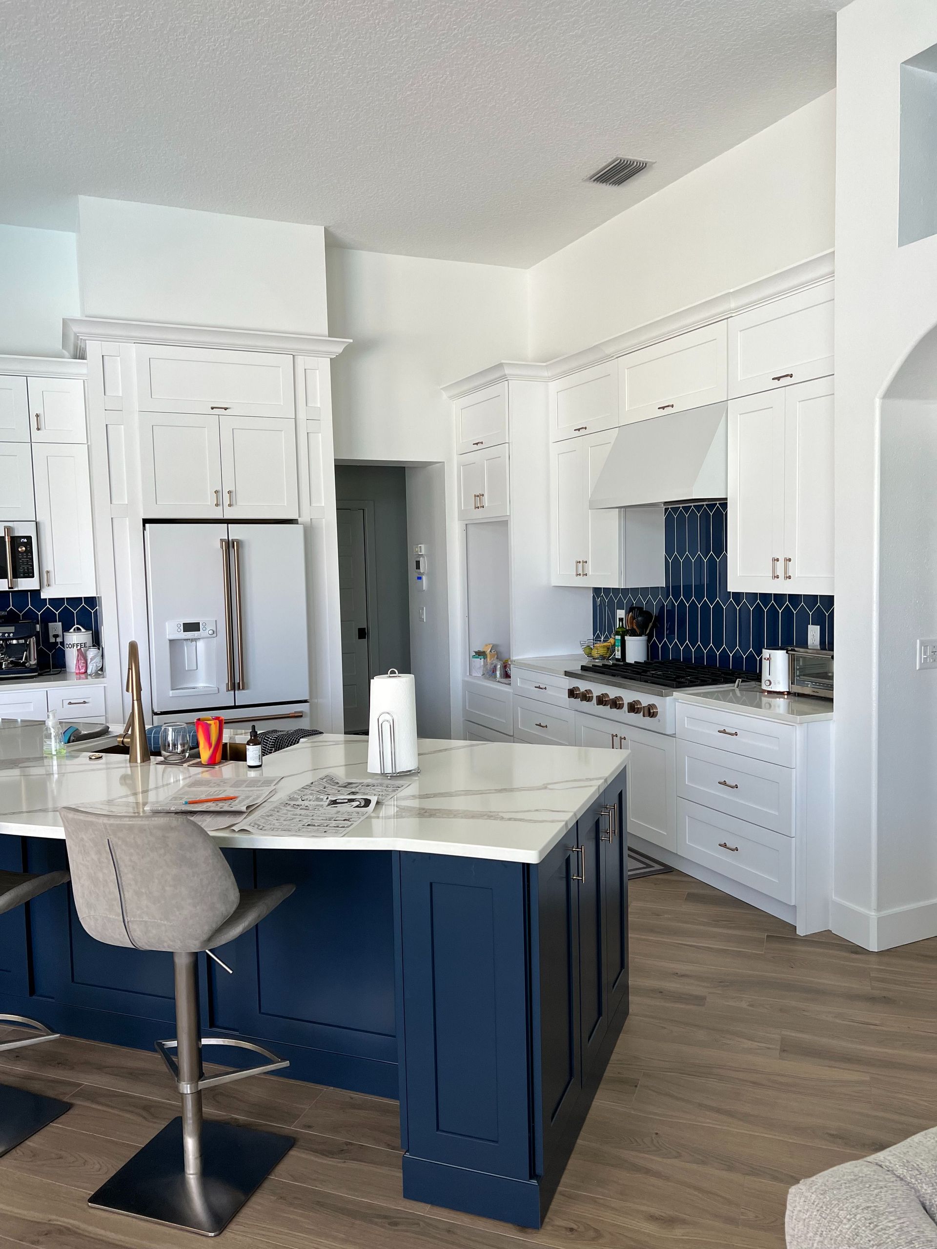 Modern white kitchen with a dark blue island, marble countertops, and blue tile backsplash on light wood floors.