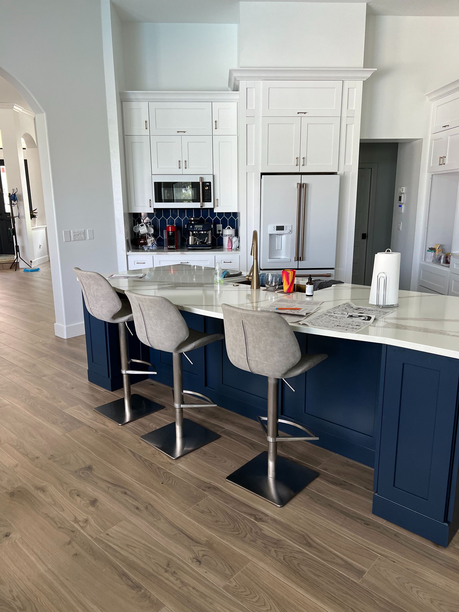 A kitchen island with three stools in front of white cabinets, a microwave, a stainless refrigerator, and wood floors.