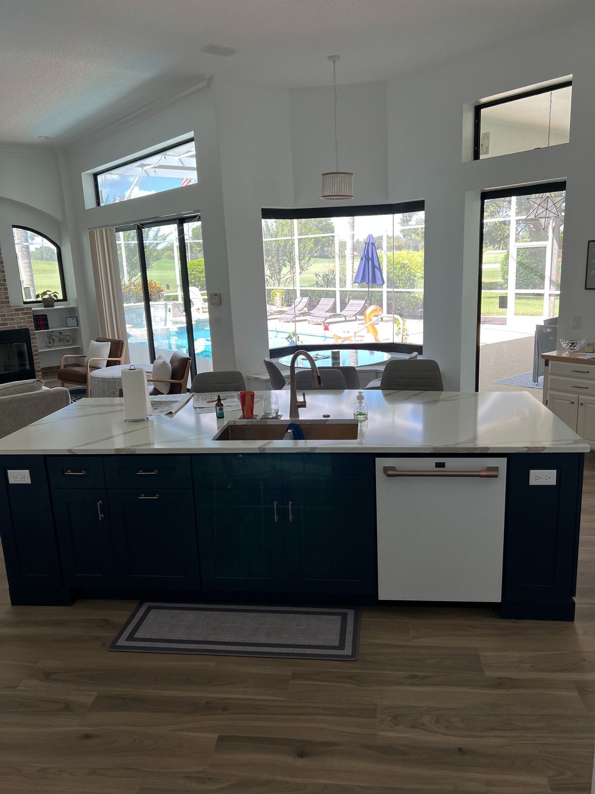 A dark blue kitchen island with a white dishwasher and marble countertop in a bright room with views of a pool area.