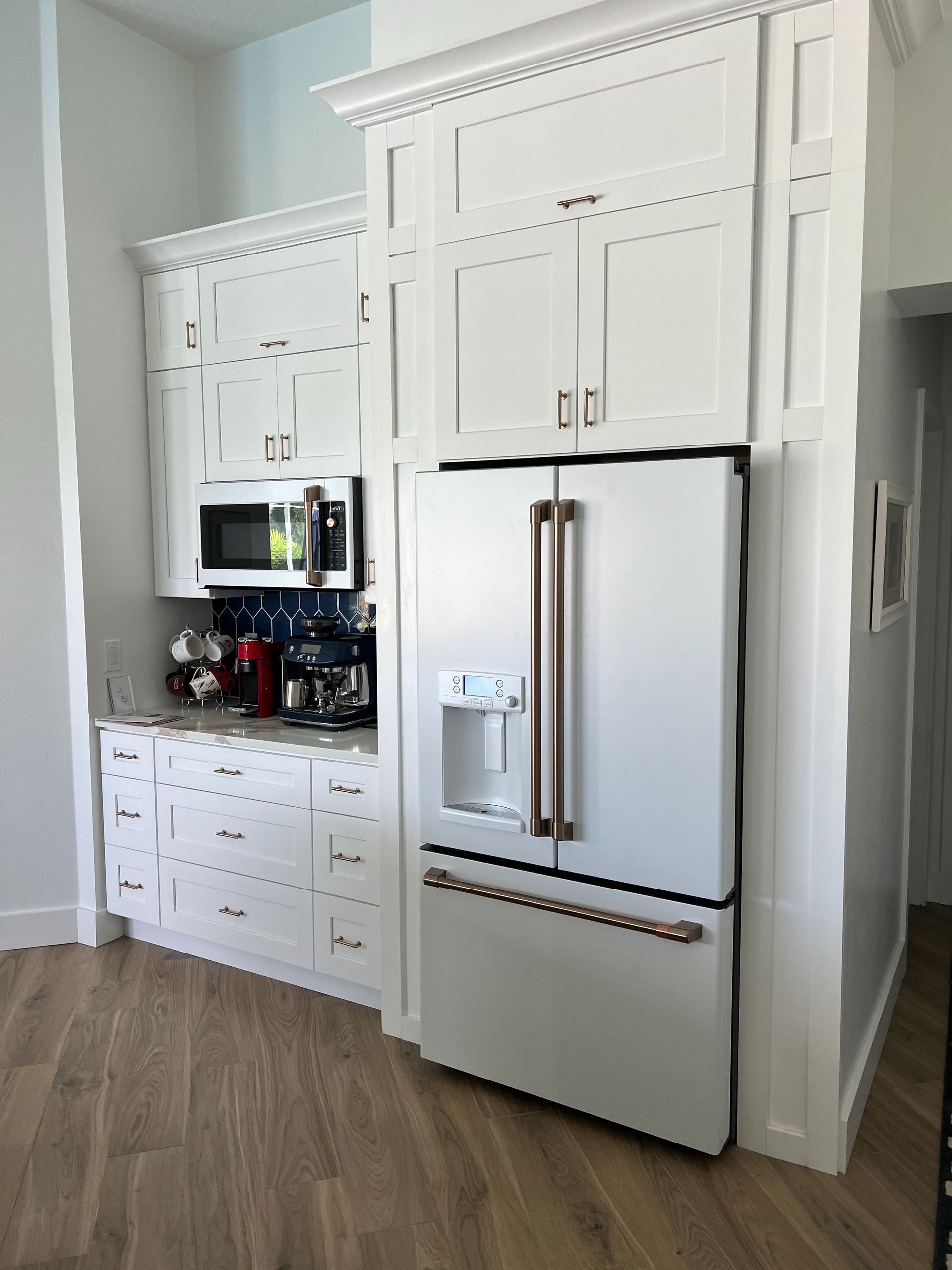 White kitchen with a stainless steel refrigerator featuring rose gold handles, next to cabinetry and a built-in microwave.