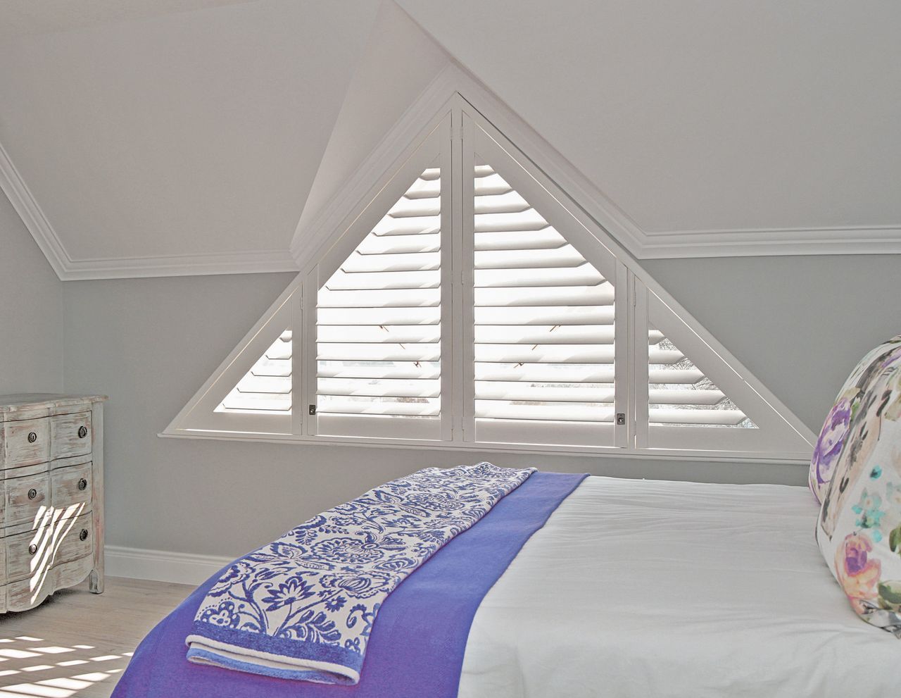 A triangular window with white shutters in a bedroom, positioned above a bed with a blue and white patterned throw.