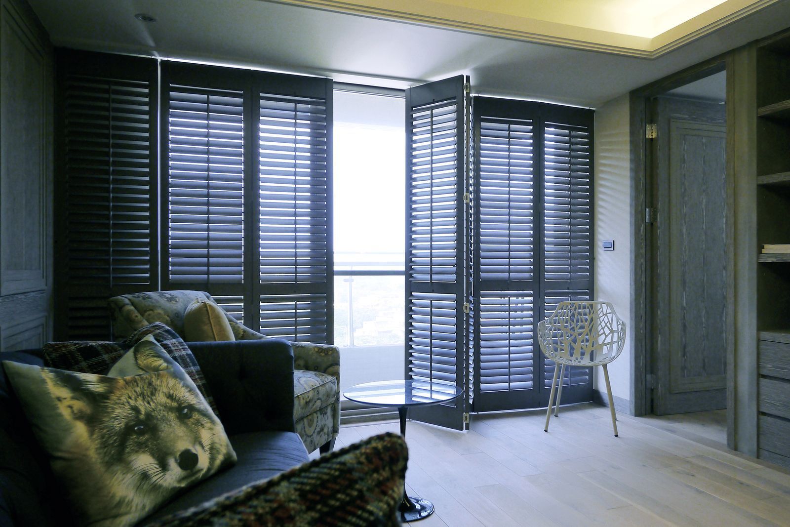 A modern room with dark wooden shutters over a bright window, a sofa with a wolf-patterned cushion, and a metal chair.
