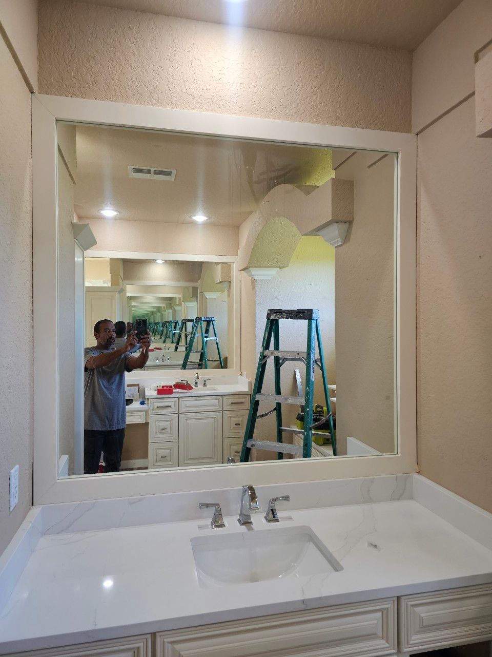 A white-framed mirror reflects a bathroom with a white vanity, countertop, and a green step ladder.