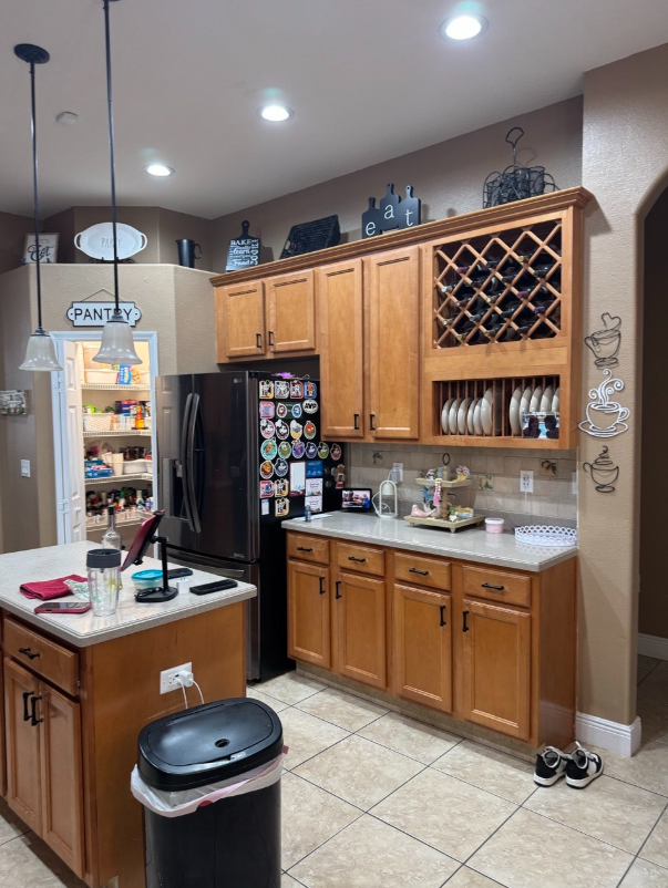A kitchen with light wood cabinets, granite countertops, a black refrigerator covered in magnets, and a pantry doorway.